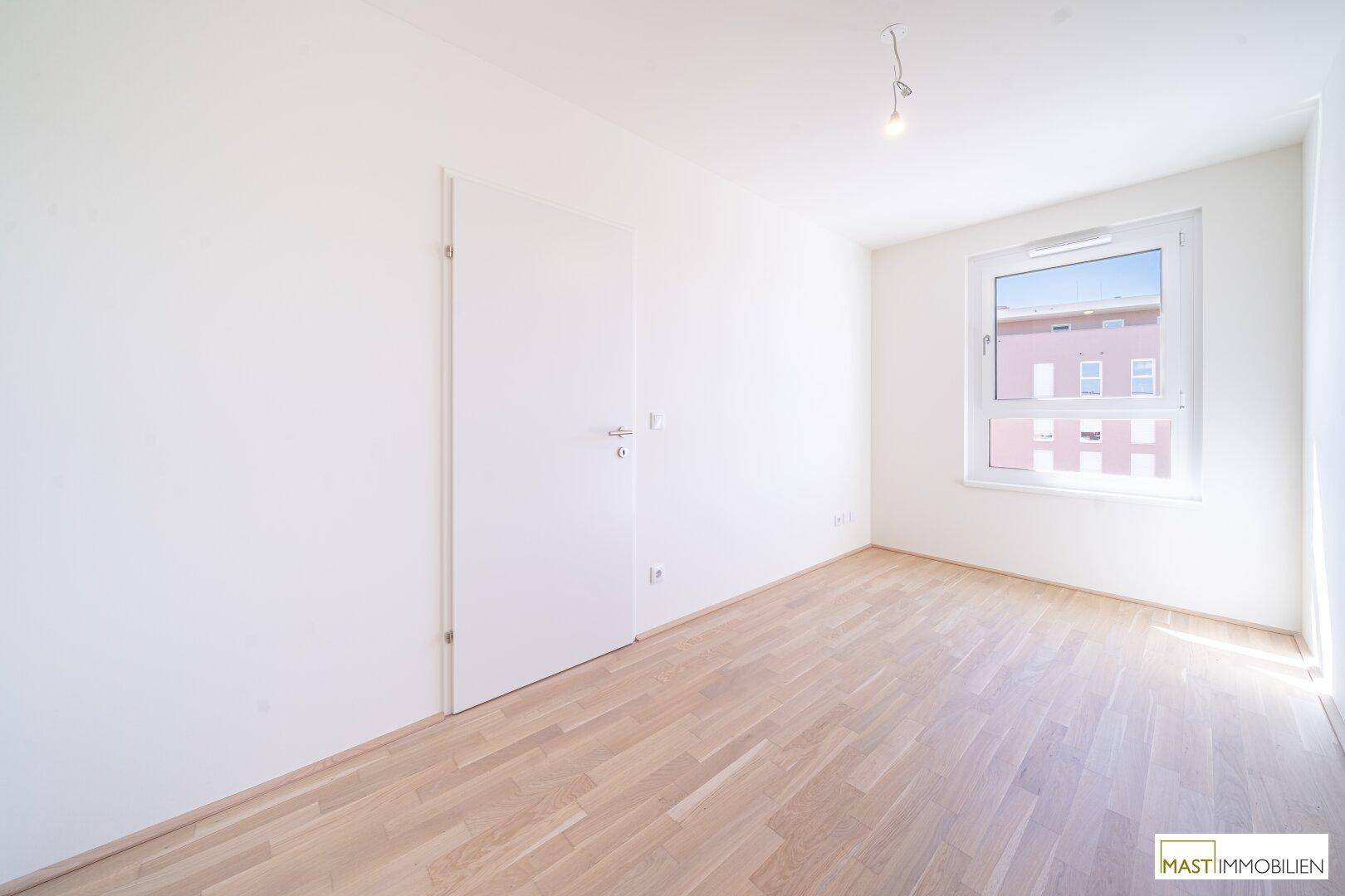 Helles Schlafzimmer mit Parkettboden und einem großen Fenster, das den Raum erhellt.