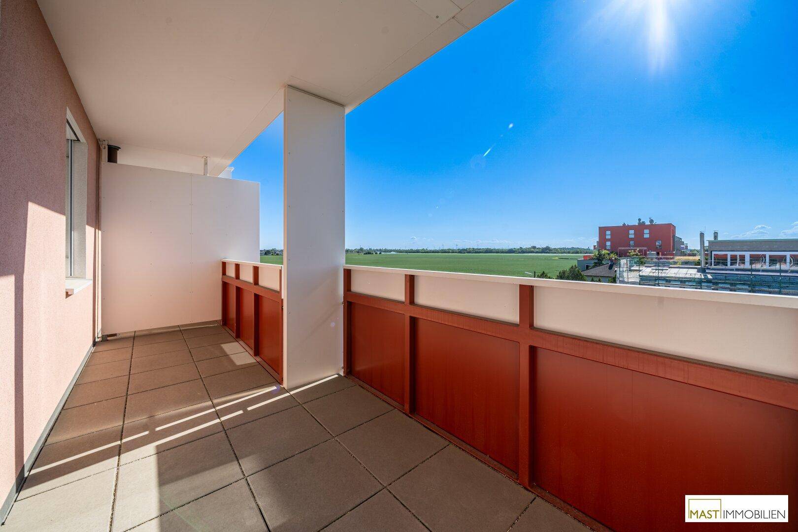 Sonniger Balkon mit Fliesenboden und rotem Geländer, bietet einen weiten Blick über grüne Felder.