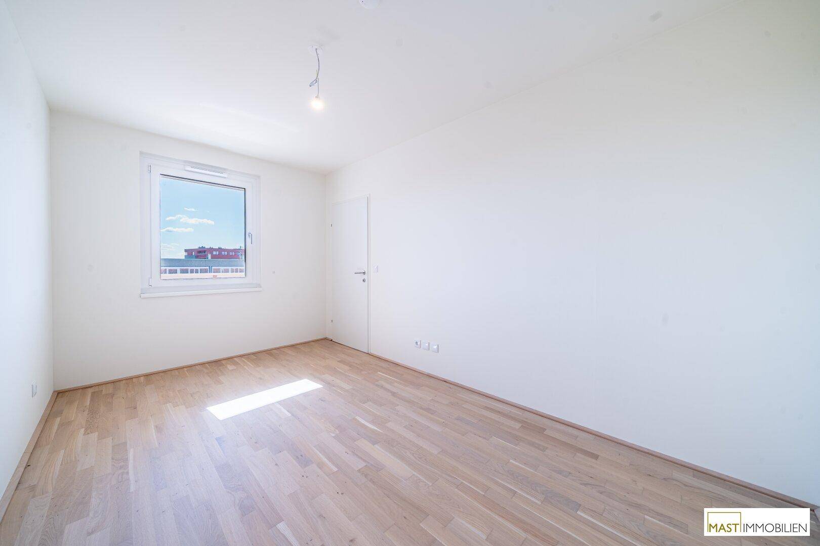 Helles Schlafzimmer mit Parkettboden und einem großen Fenster, das viel Tageslicht hereinlässt.