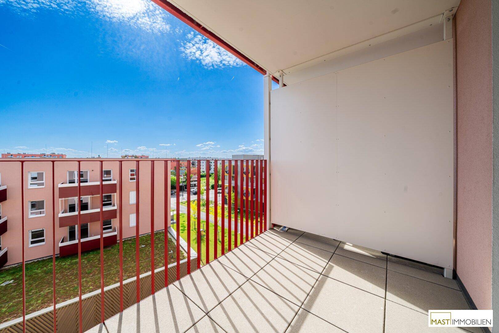 Sonniger Balkon mit Fliesenboden, rotem Geländer und seitlichem Sichtschutz, mit Blick auf die Umgebung.