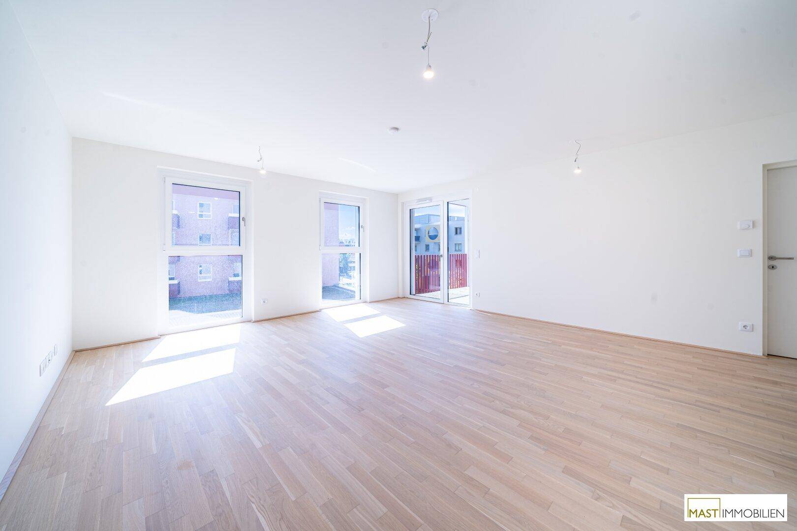 Helles, leeres Wohnzimmer mit Parkettboden und Zugang zu einem Balkon, durchflutet von Sonnenlicht.