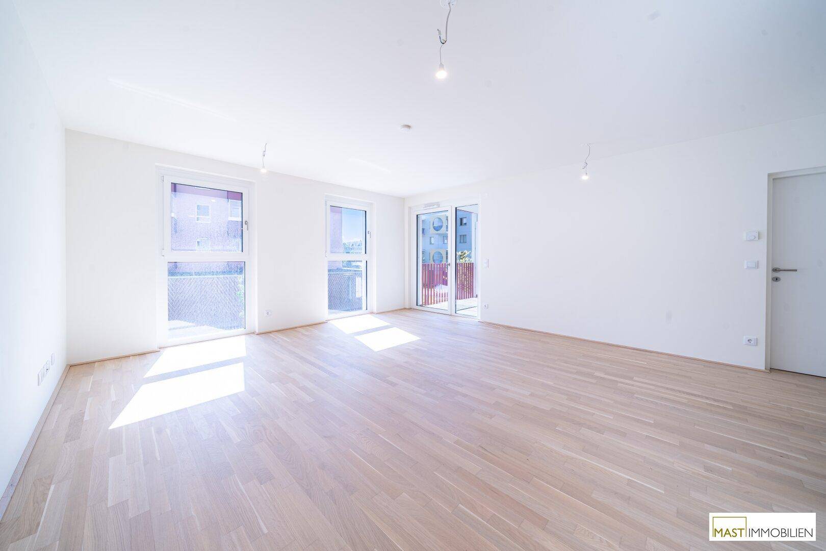 Heller, leerer Wohnraum mit hellem Parkettboden, großen Fenstern und Zugang zu einem Balkon.