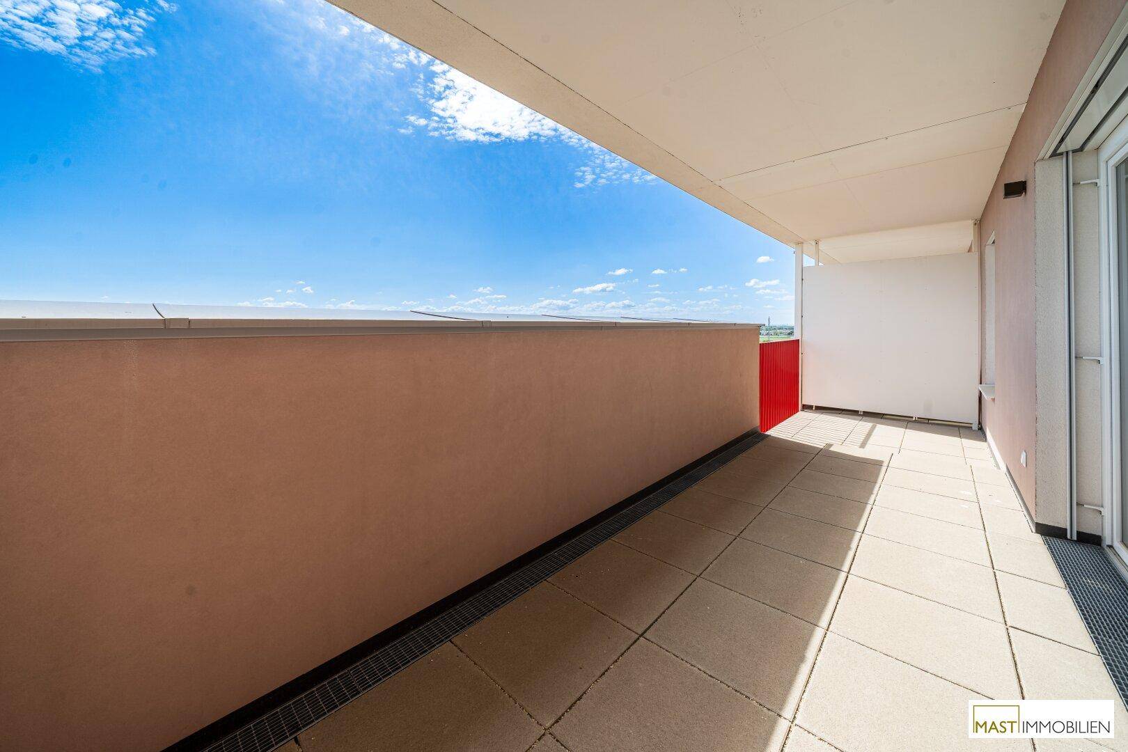 Lange, sonnige Terrasse mit Fliesenboden und Blick auf die umliegenden Gebäude.