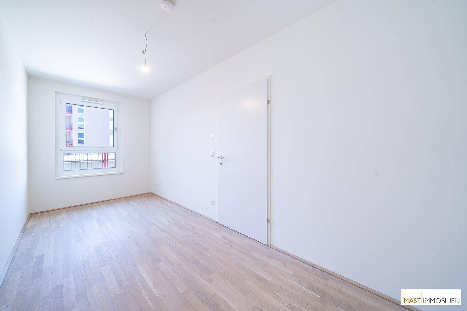 Leeres Schlafzimmer mit hellem Parkettboden und einem Fenster mit Blick nach draußen.