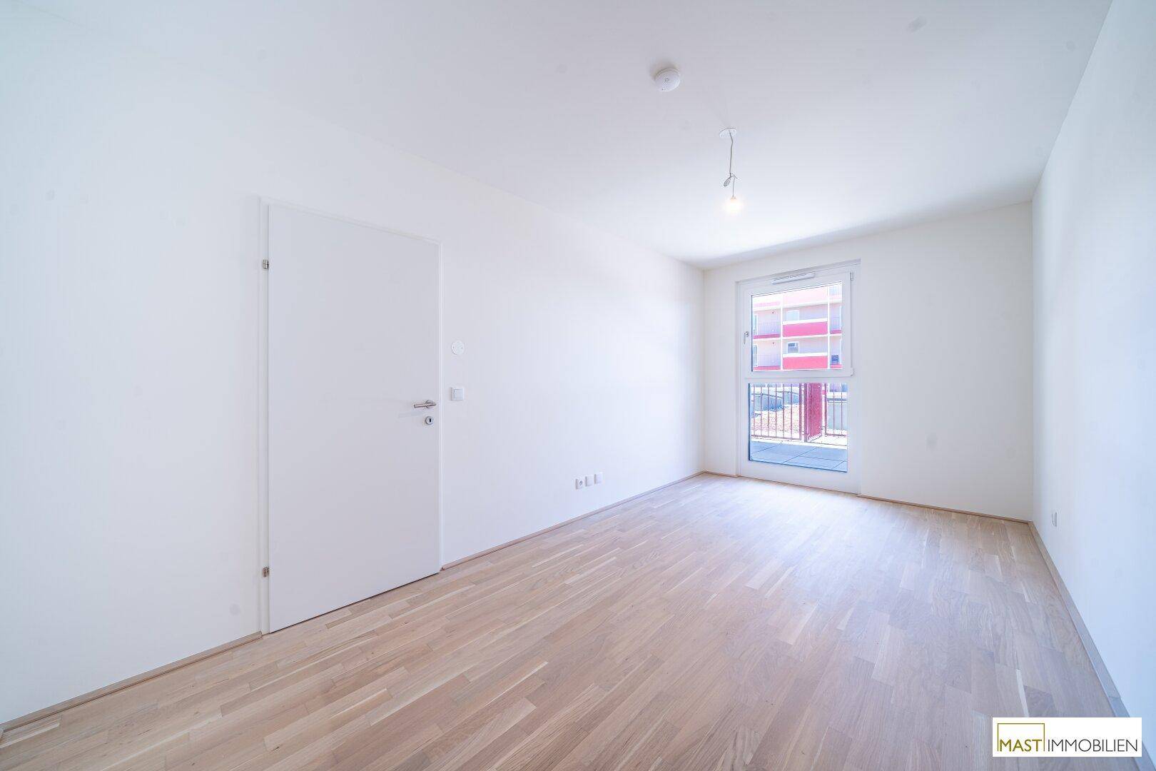 Helles Zimmer mit Parkettboden und großem Fenster mit Zugang zu einem Außenbereich.