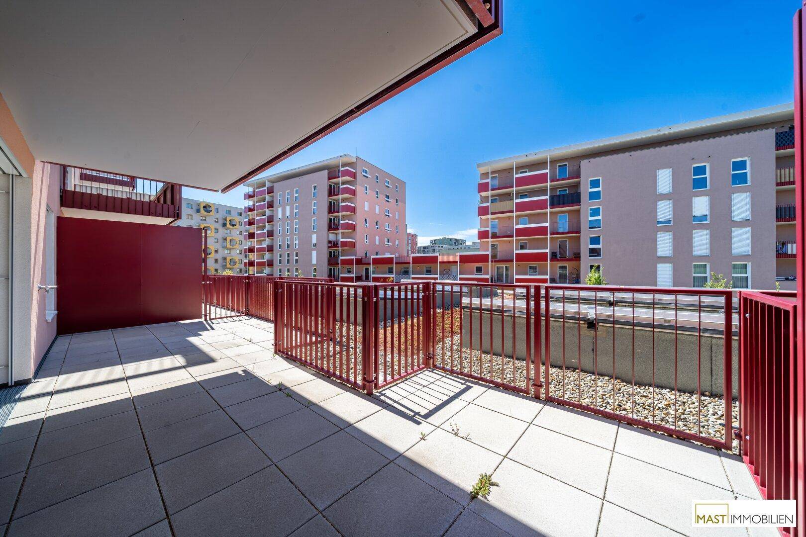 Sonnige Terrasse mit roten Geländern und Blick auf die umliegenden Wohngebäude.
