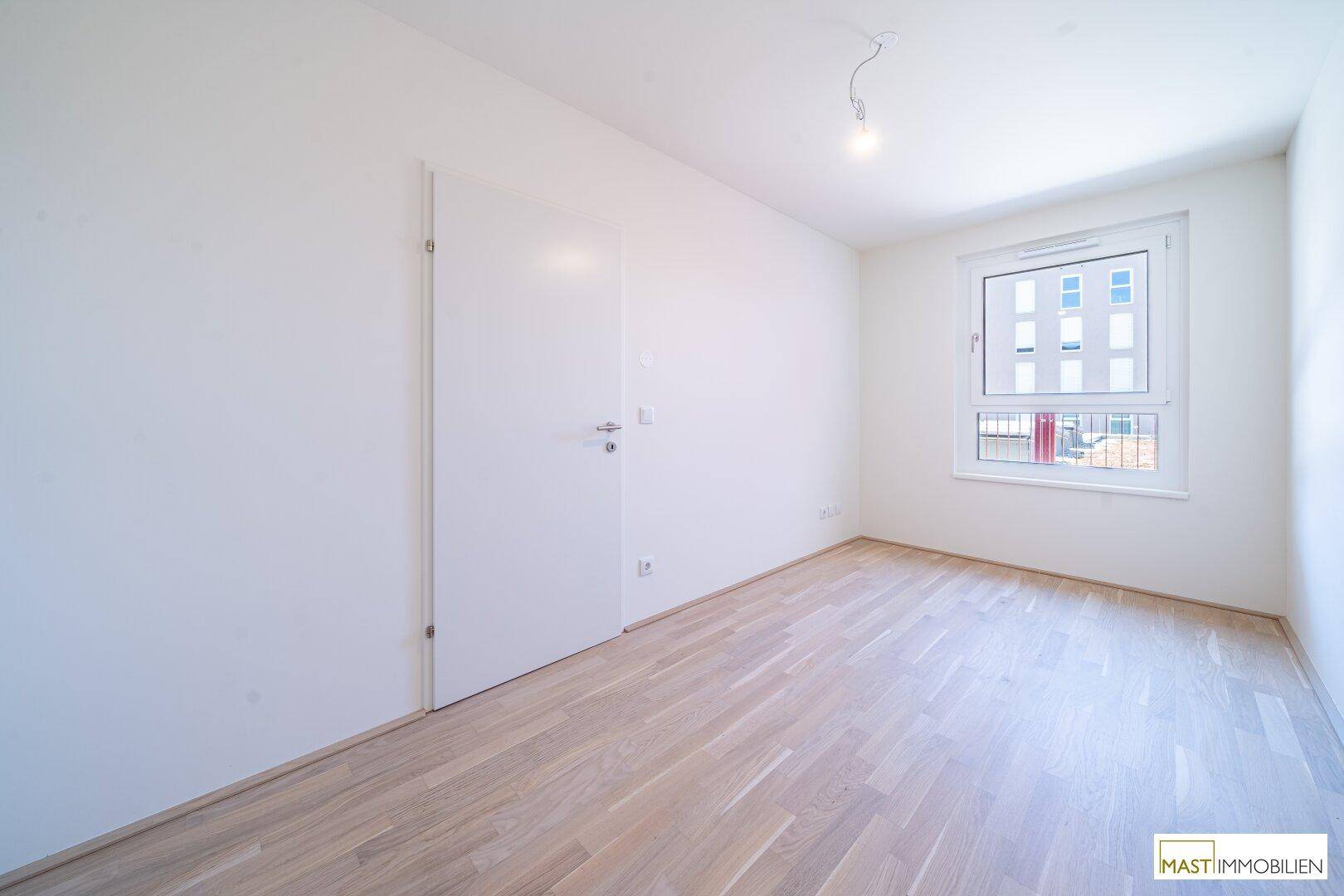 Leeres Zimmer mit Holzboden und großem Fenster, ideal als Schlaf- oder Arbeitszimmer nutzbar.