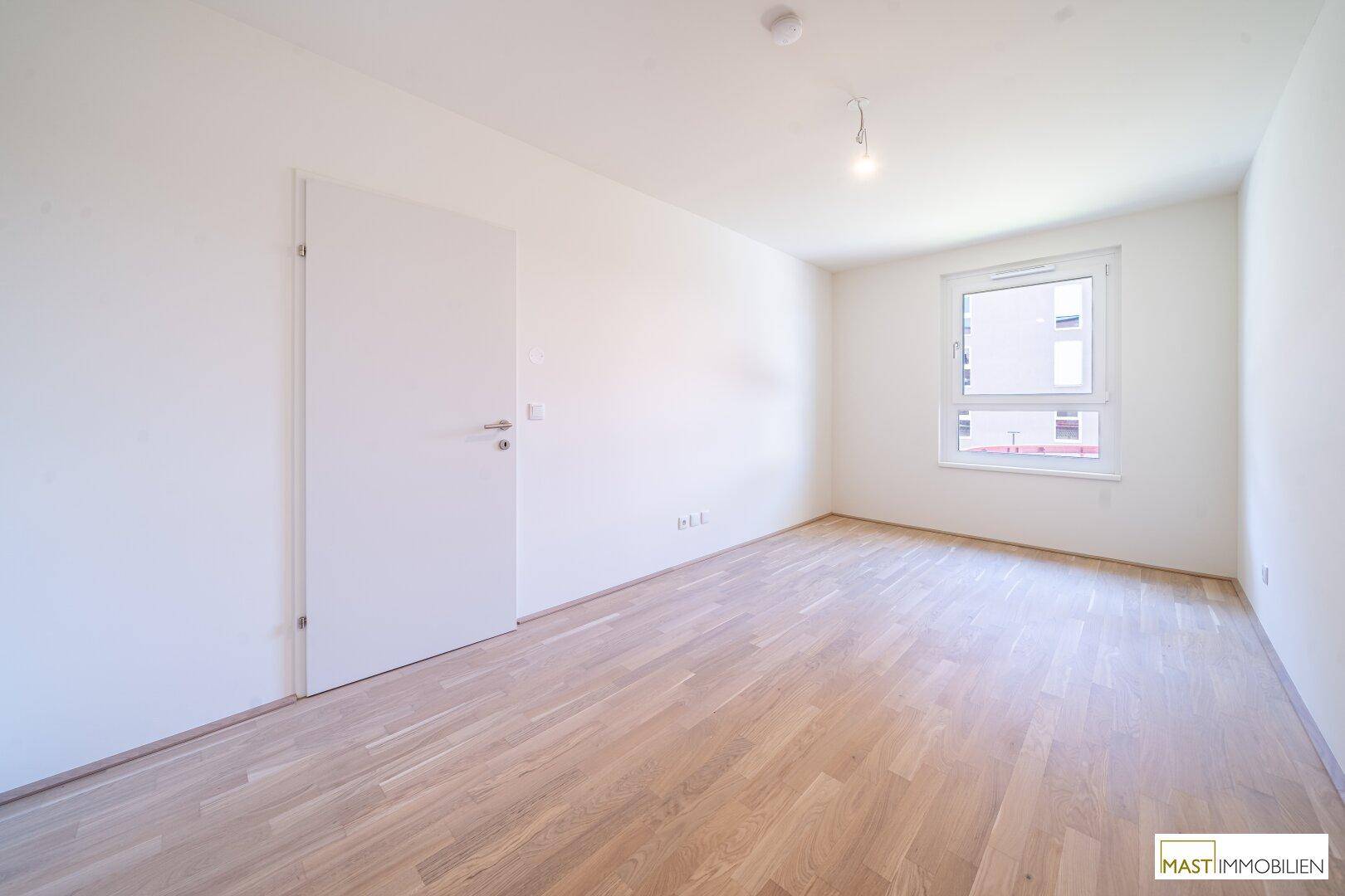 Helles Schlafzimmer mit Parkettboden und einem großen Fenster, das viel Tageslicht hereinlässt.