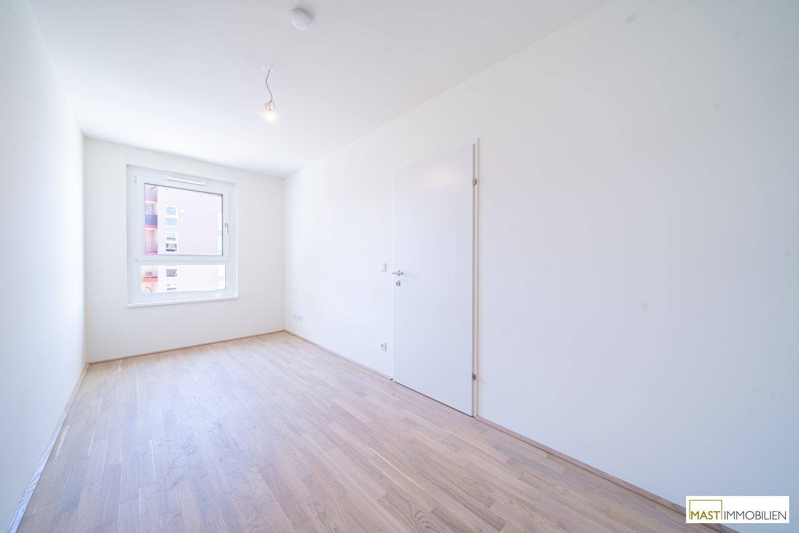 Freundliches, leeres Zimmer mit hellem Holzparkettboden und großem Fenster für viel Tageslicht.