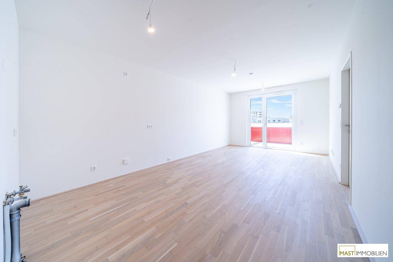 Heller, leerer Wohnraum mit weiß gestrichenen Wänden und hellem Holzparkettboden, mit Zugang zum Balkon.