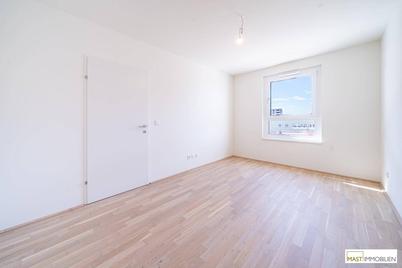 Leeres Schlafzimmer mit hellen Holzböden und einem großen Fenster, das viel Tageslicht hereinlässt.