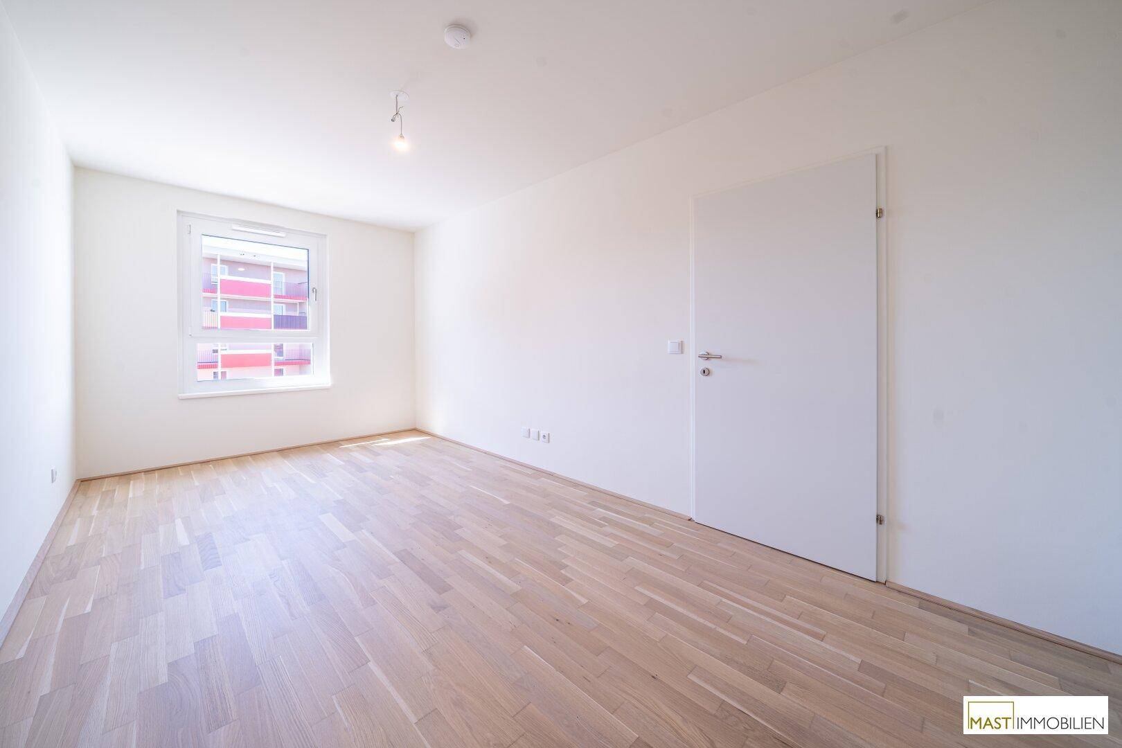 Leeres Schlafzimmer mit hellem Holzfußboden und einem Fenster, das viel Tageslicht hereinlässt.