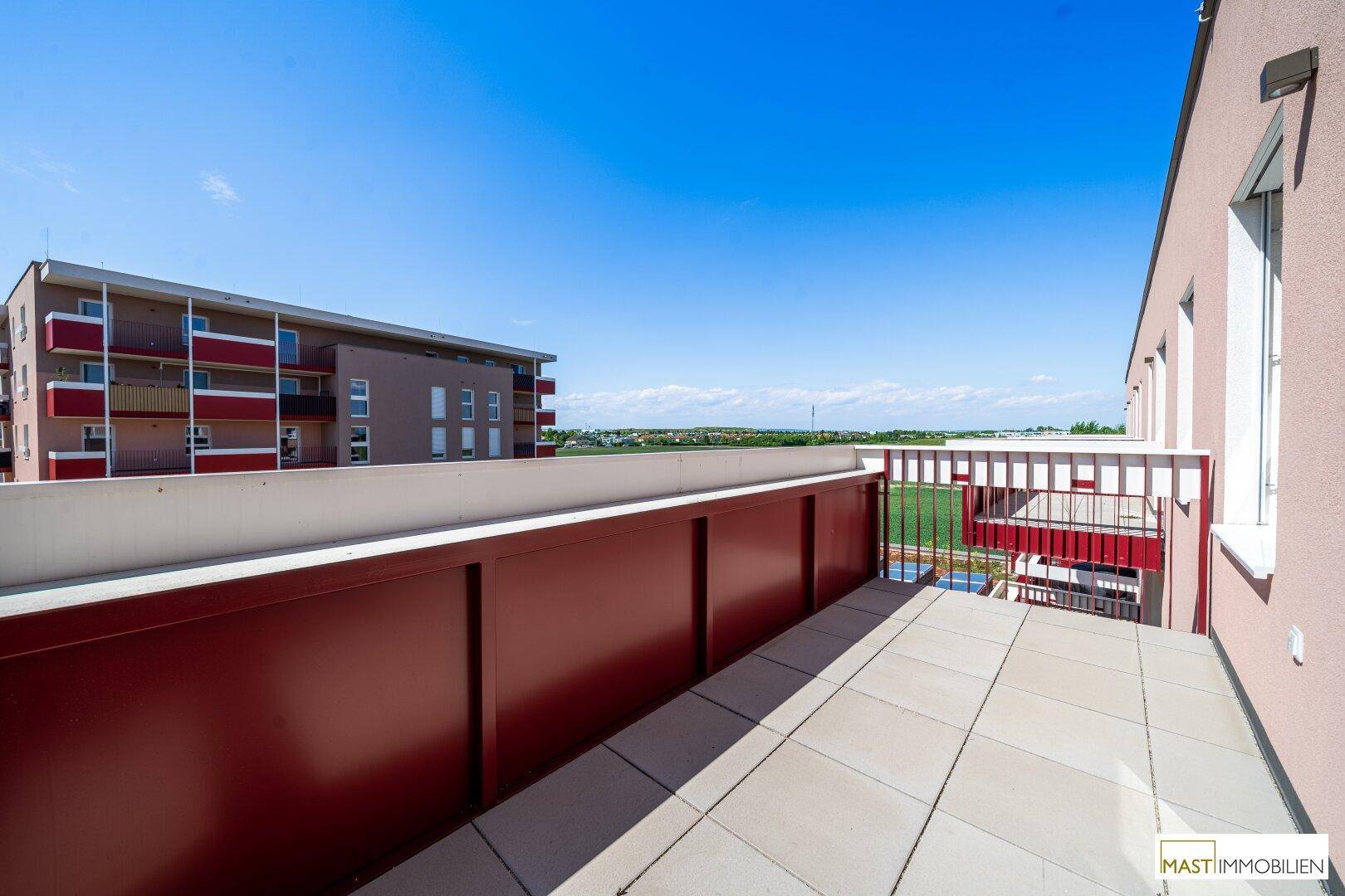 Sonnige Terrasse mit Fliesenboden und rotem Geländer, bietet einen Blick auf die Umgebung und andere Gebäude.