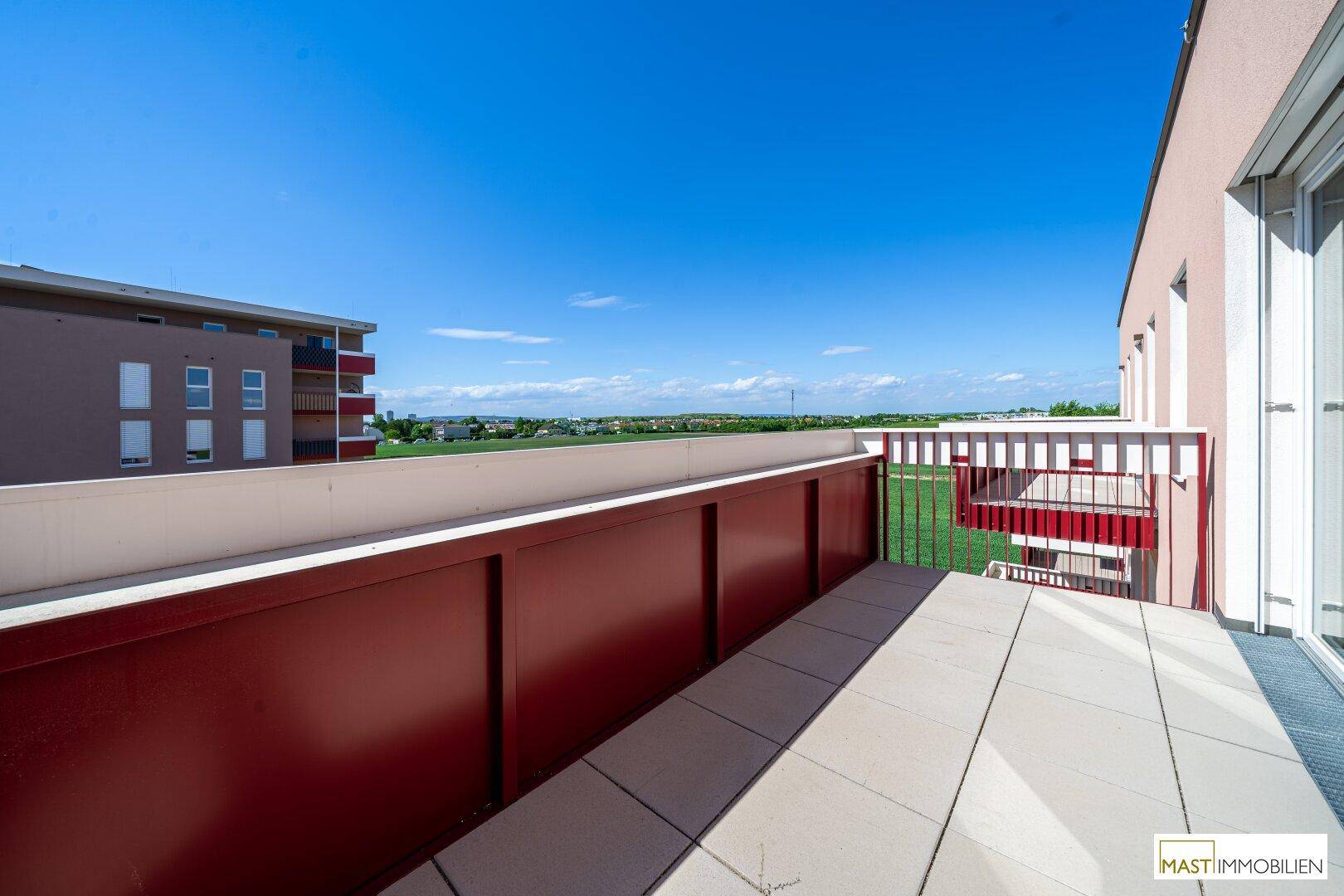 Geräumiger Balkon mit rotem Geländer und weitem Blick über die Umgebung und grüne Felder.