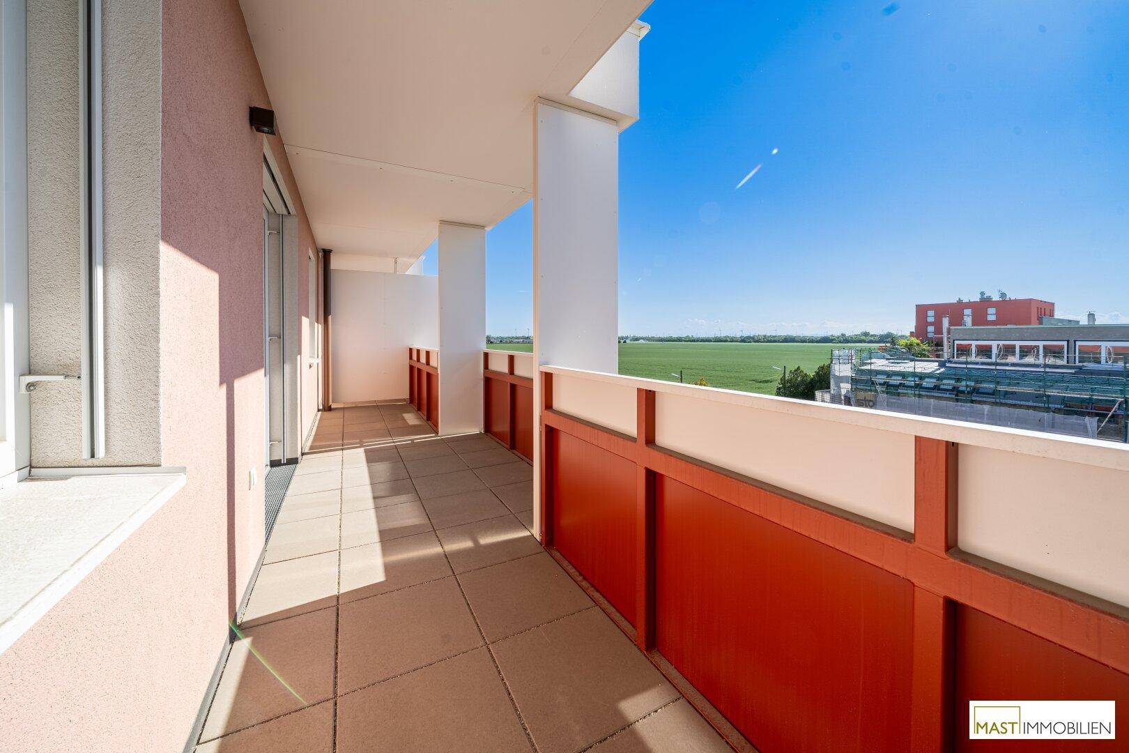 Geräumiger Balkon mit Fliesenboden, rotem Geländer und weitem Blick ins Grüne.