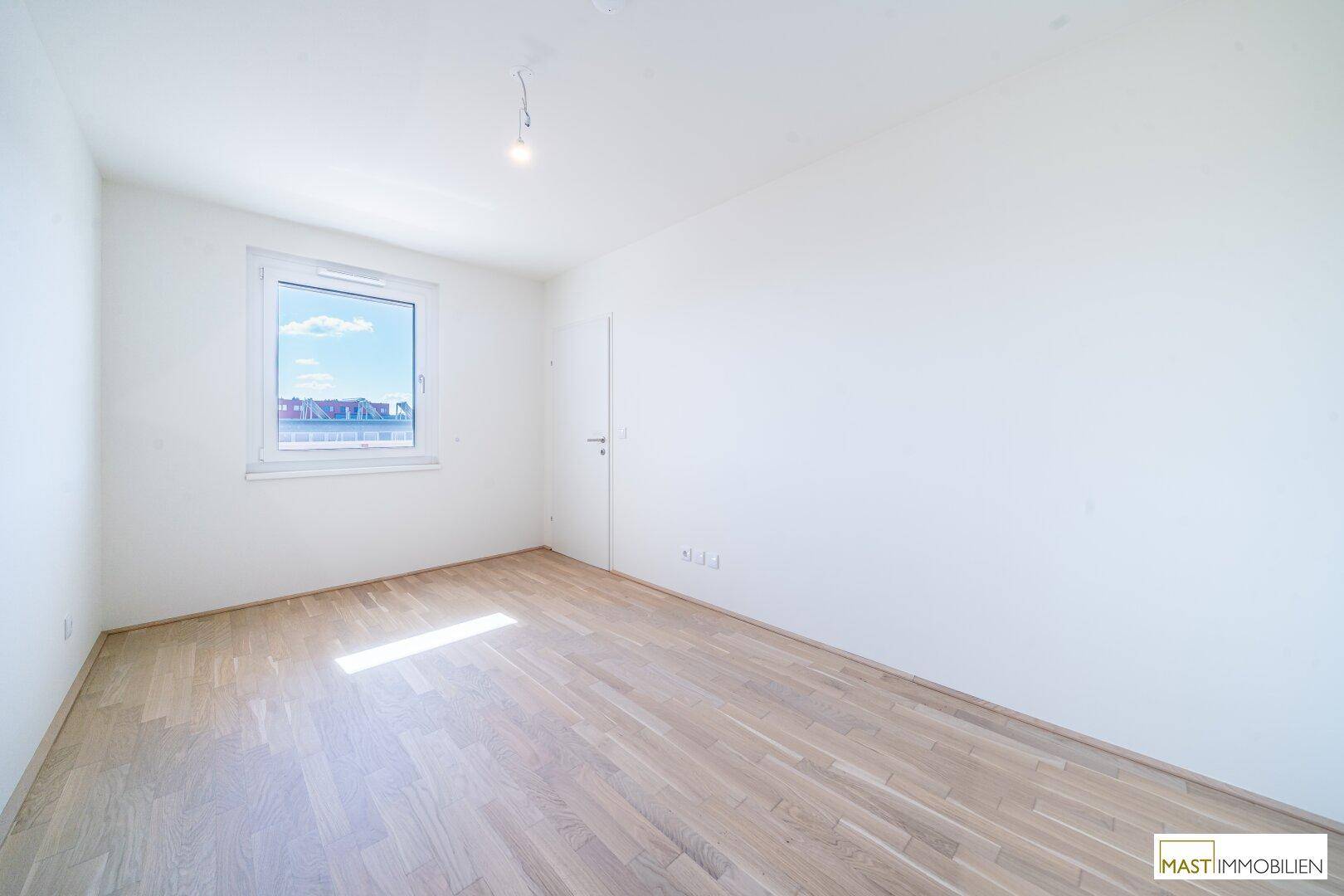 Helles Schlafzimmer mit Holzparkettboden und einem großen Fenster, das viel Tageslicht hereinlässt.