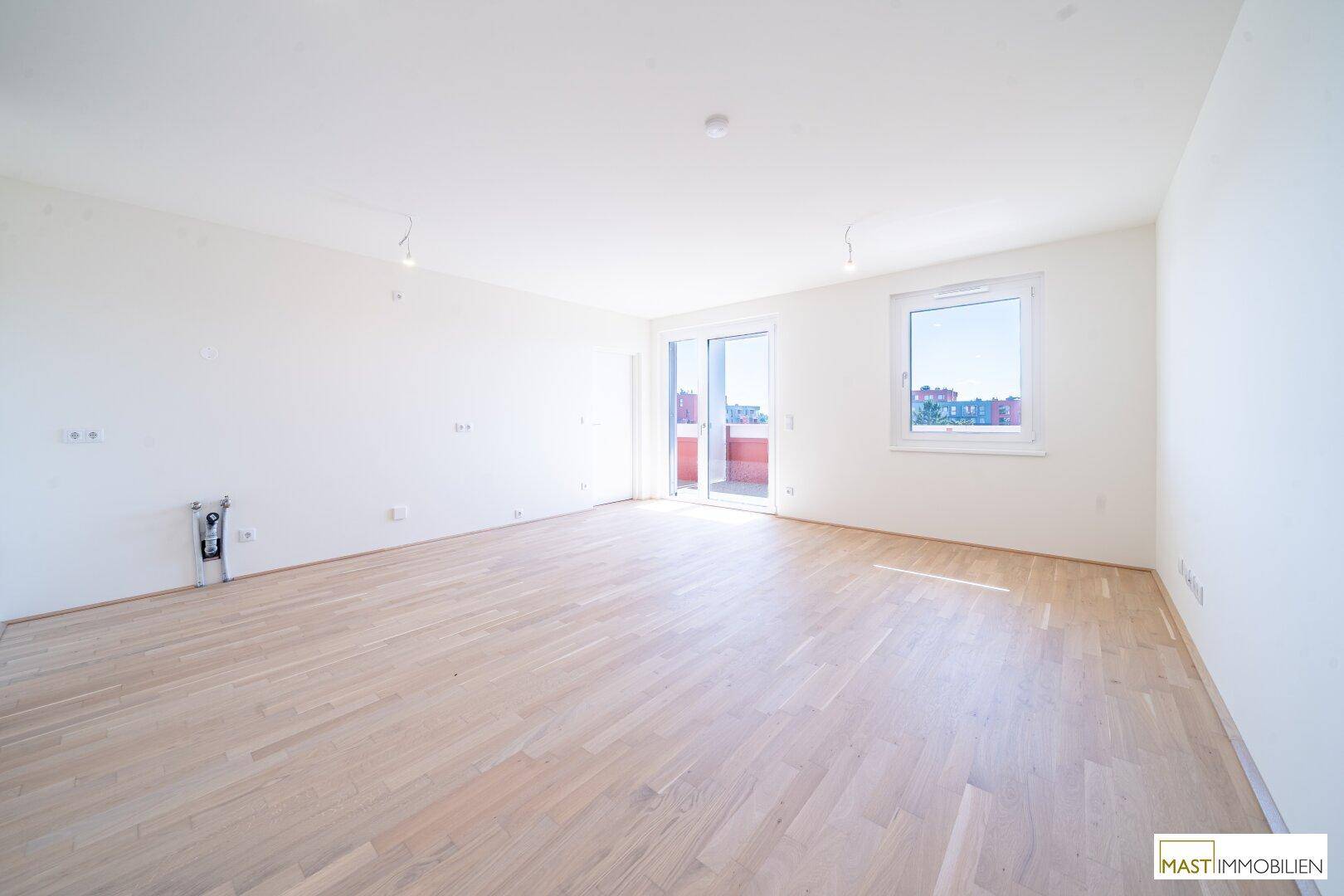 Leeres Wohnzimmer mit hellem Parkettboden, großen Fenstern und Zugang zum Balkon.