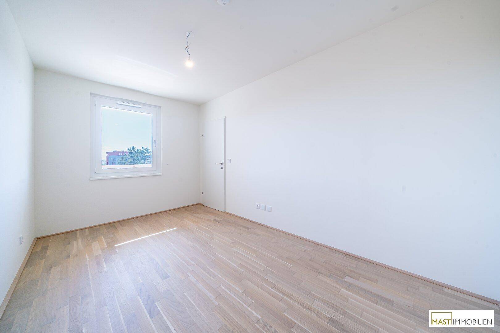 Helles, leeres Schlafzimmer mit Parkettboden und einem großen Fenster.