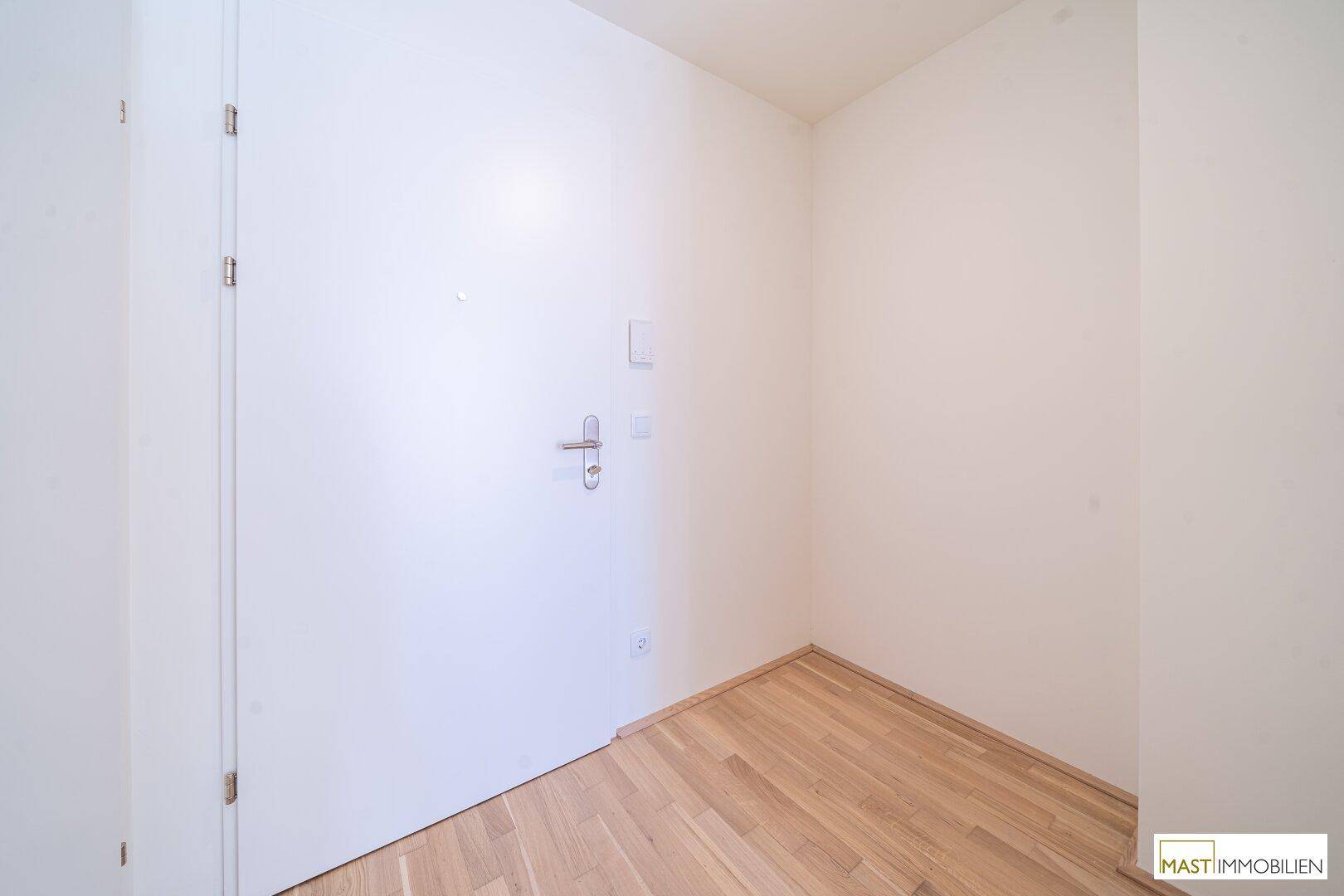 Heller Eingangsbereich mit weißer Tür und neuem Holzparkettboden in einem modernen Gebäude.