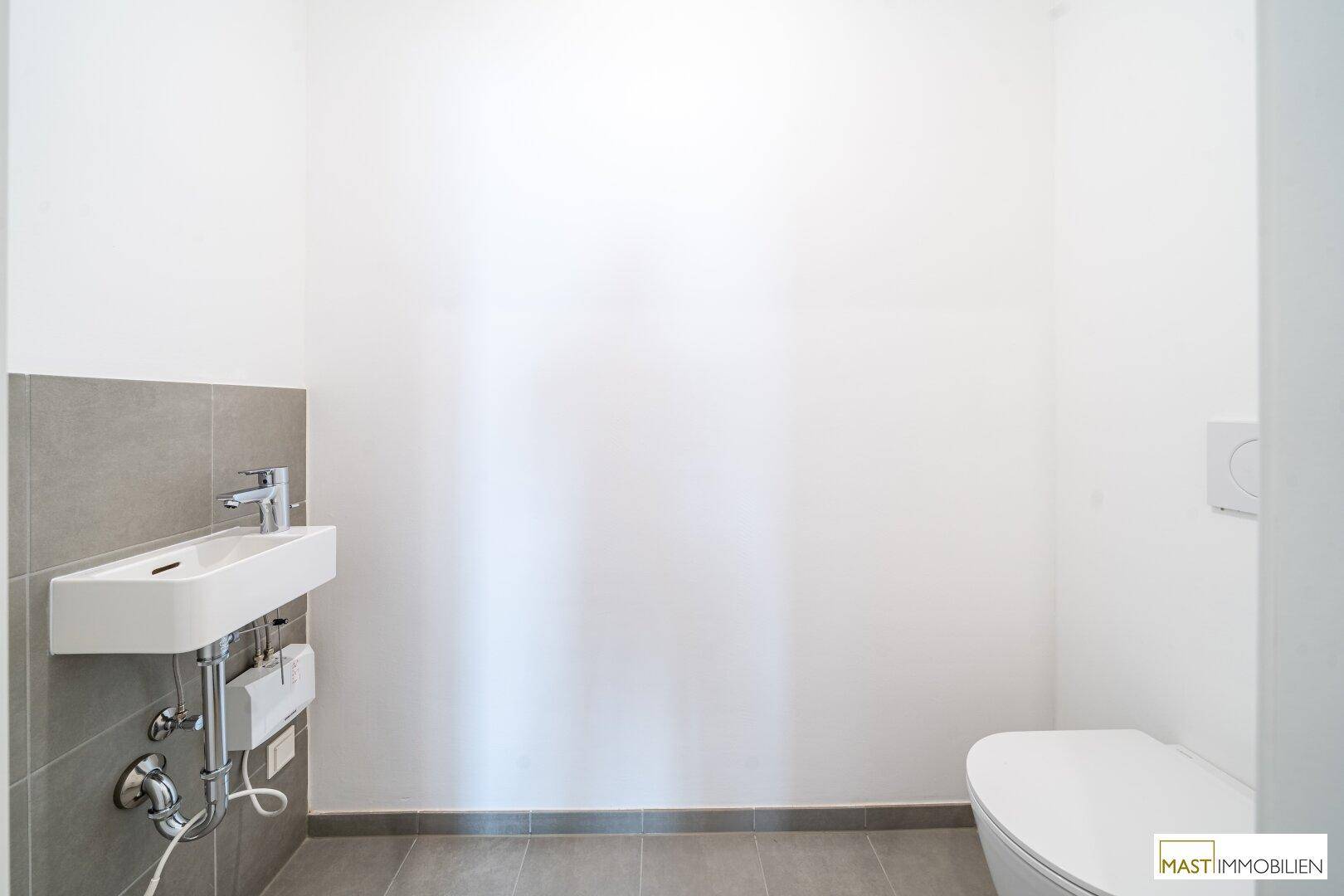 Separates WC mit Toilette und kleinem Waschbecken, teilweise mit grauen Fliesen verkleidet.