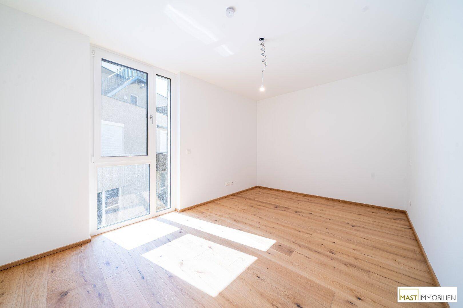 Leeres Schlafzimmer mit großem Fenster, hellem Holzboden und viel Tageslicht.