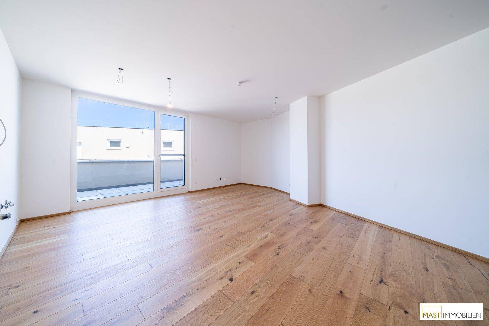 Heller, leerer Wohnraum mit großen Fenstern und neuem Holzparkettboden.