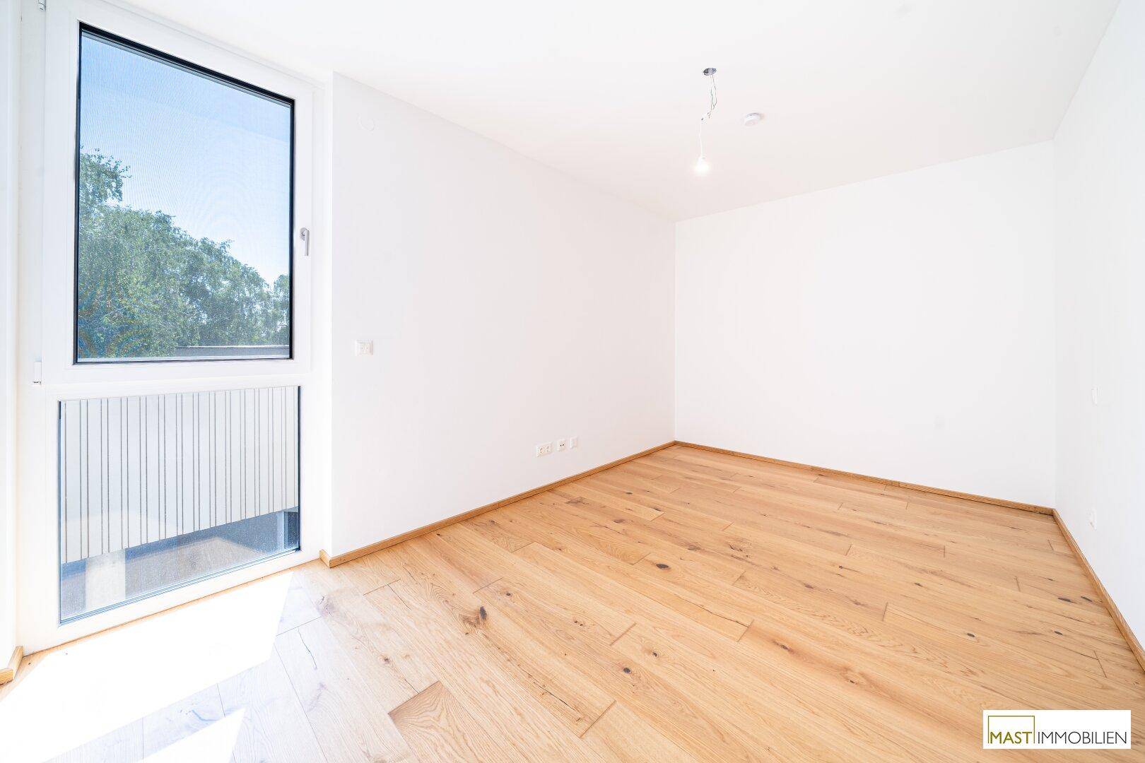 Unmöbliertes Zimmer mit großem Fenster und hellem Holzparkettboden, ideal als Schlafzimmer.
