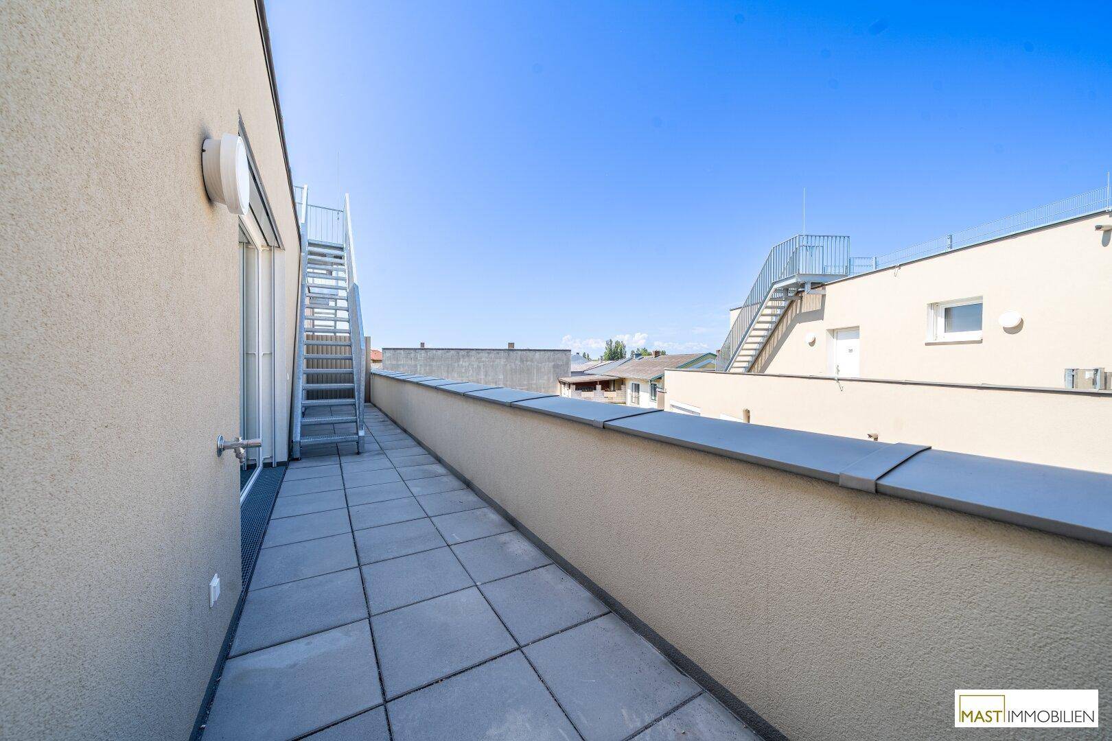 Langer Balkon mit Fliesenboden und Metallgeländer, führt zu einer Dachterrasse.