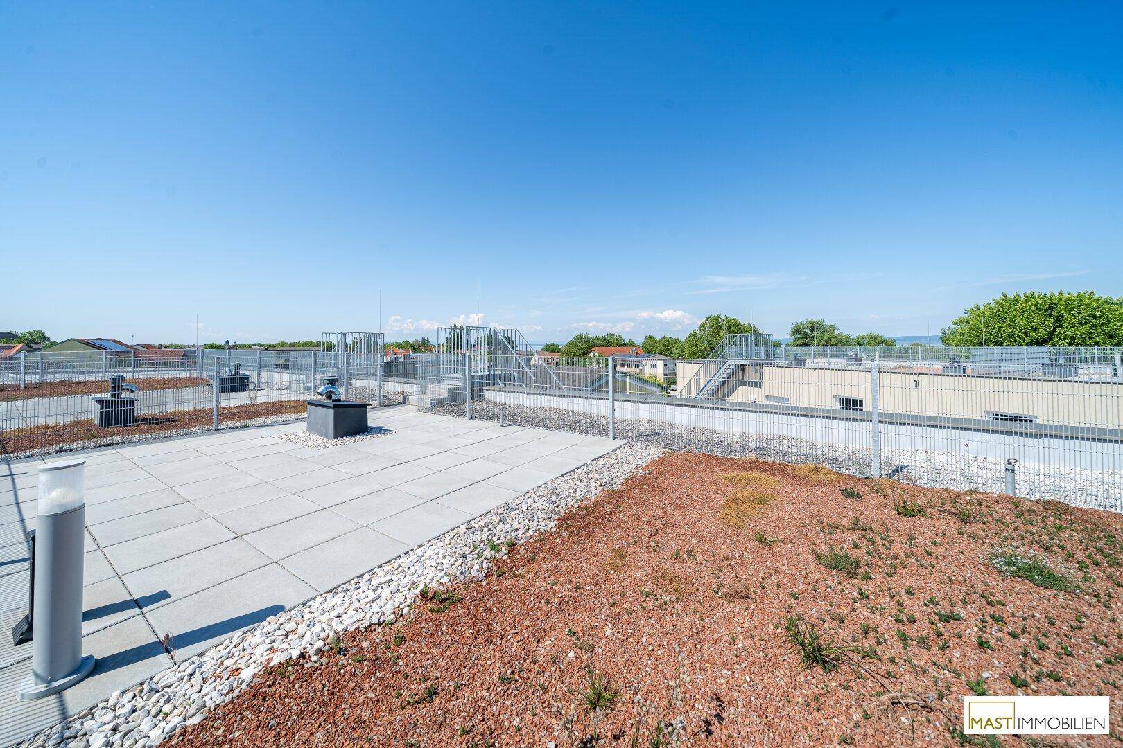 Weitläufige Dachterrasse mit Fliesen und Kiesbereichen unter klarem blauem Himmel.