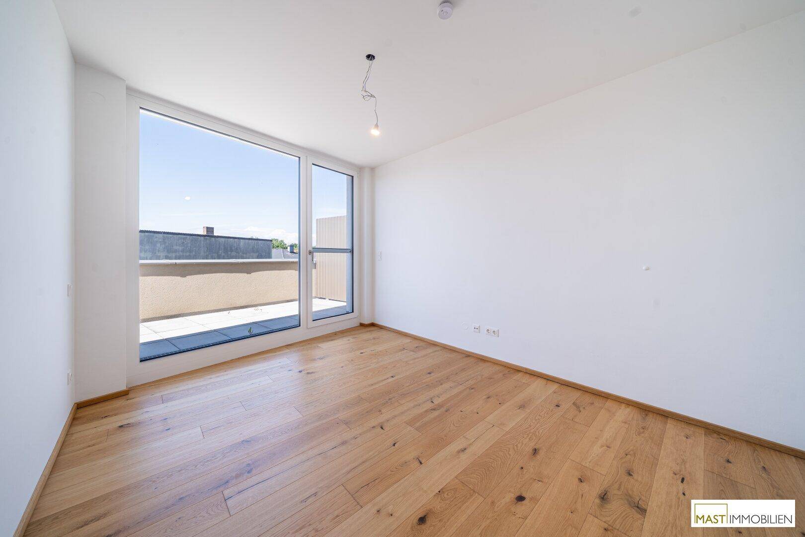 Helles Zimmer mit Holzboden und großen Fenstern, die einen Blick auf die Dachterrasse bieten.