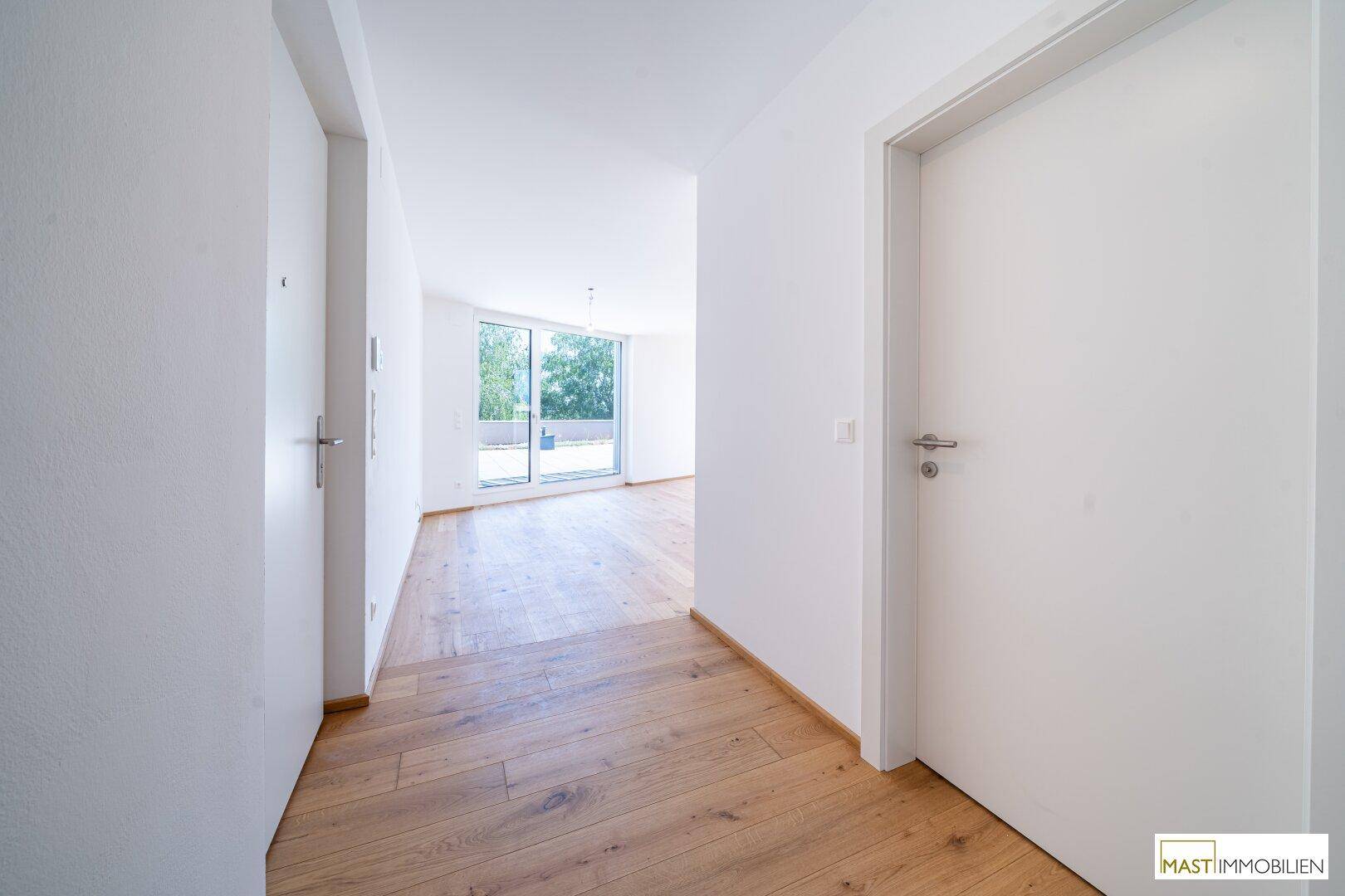 Heller Flur mit Holzboden, der zu einem lichtdurchfluteten Wohnbereich mit großen Fenstern führt.