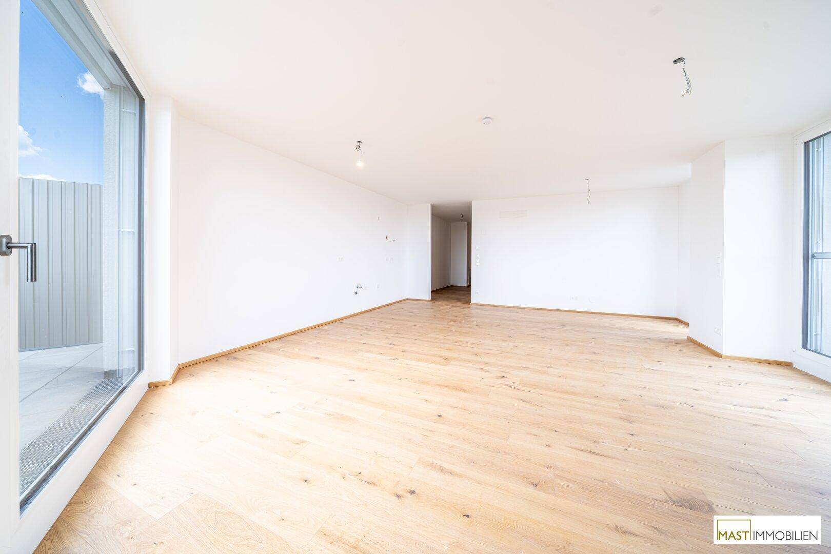 Heller, leerer Raum mit Holzboden und großem Fenster, das viel Tageslicht hereinlässt.