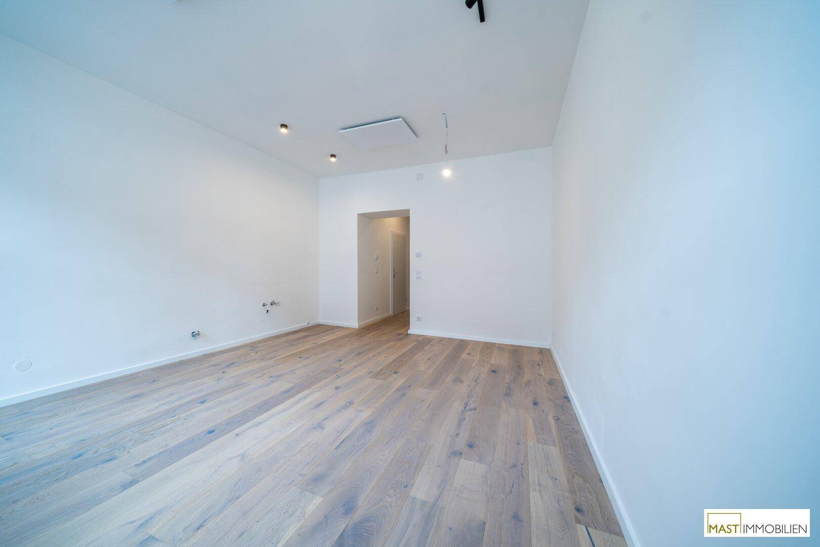 Leerer Raum mit Holzboden und weißen Wänden, mit Anschlüssen für eine mögliche Kücheninstallation.