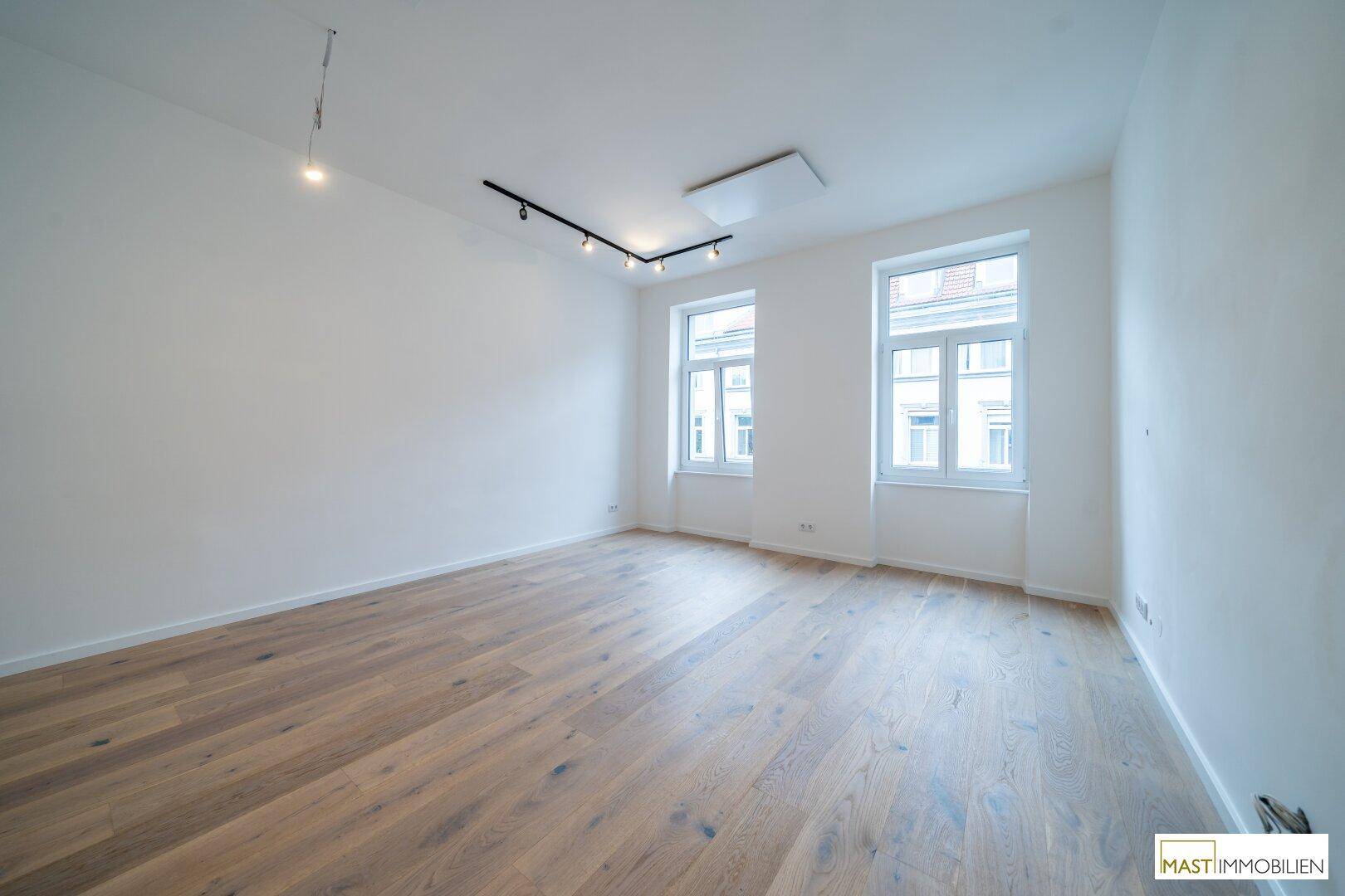 Lichtdurchfluteter Raum mit elegantem Holzboden und zwei großen Fenstern, bereit zur individuellen Gestaltung.