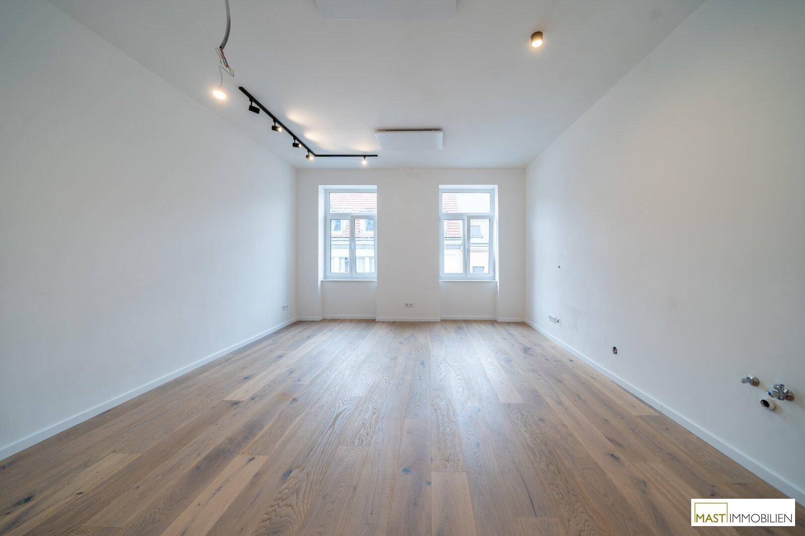 Heller Raum mit neuem Holzfußboden, weißen Wänden und zwei Fenstern, ideal als Wohnzimmer.