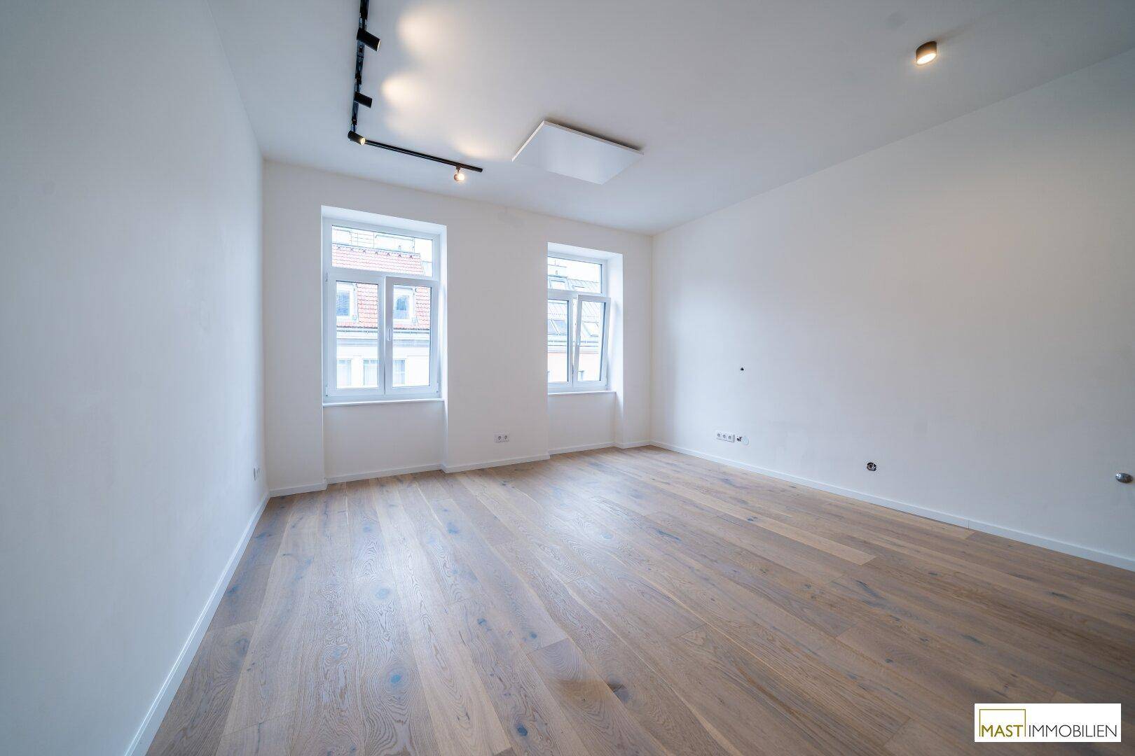 Heller Raum mit zwei Fenstern, neuem Parkettboden und moderner Schienenbeleuchtung an der Decke.