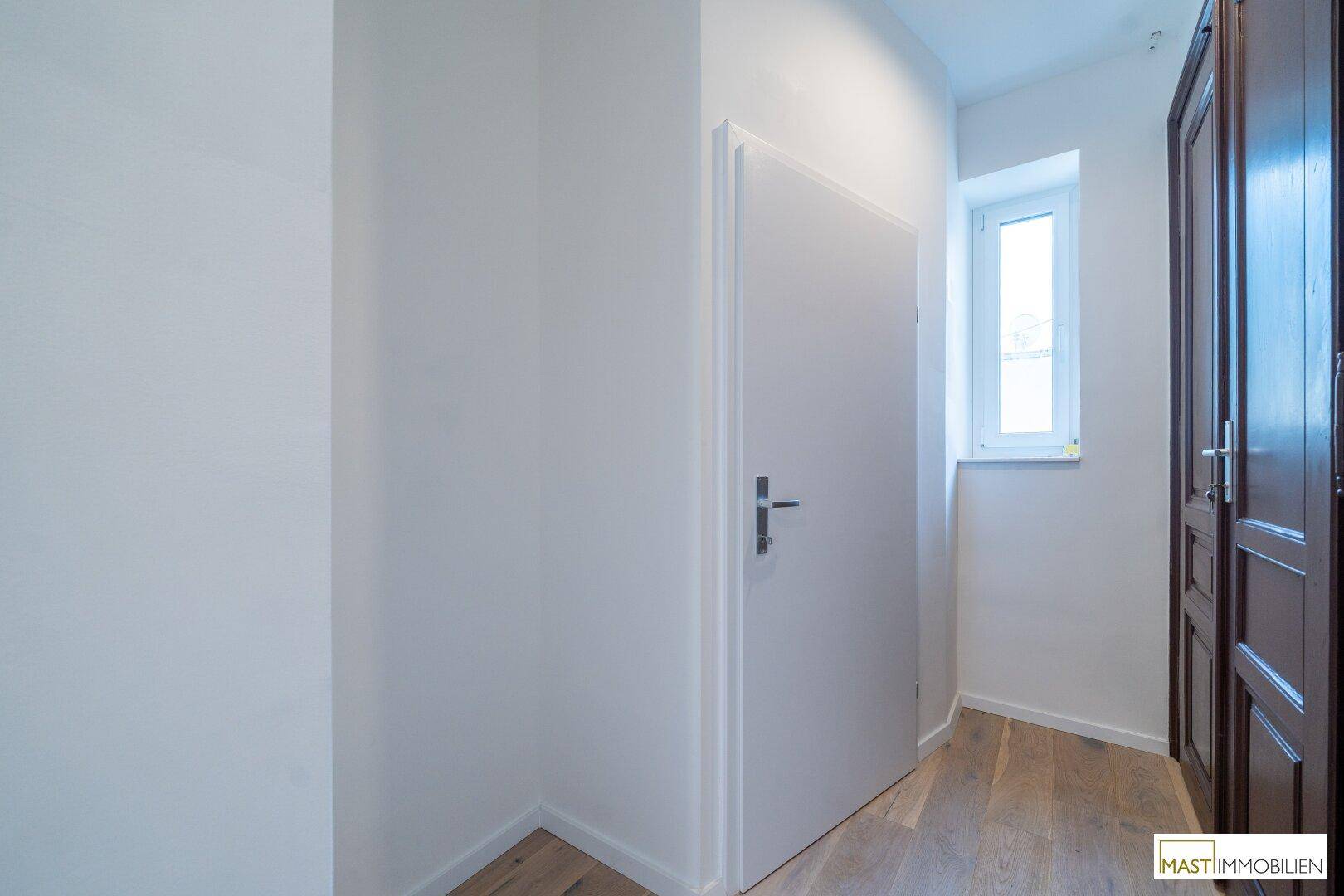 Schmaler Flur mit Holzboden, weißer Tür und einem kleinen Fenster am Ende.