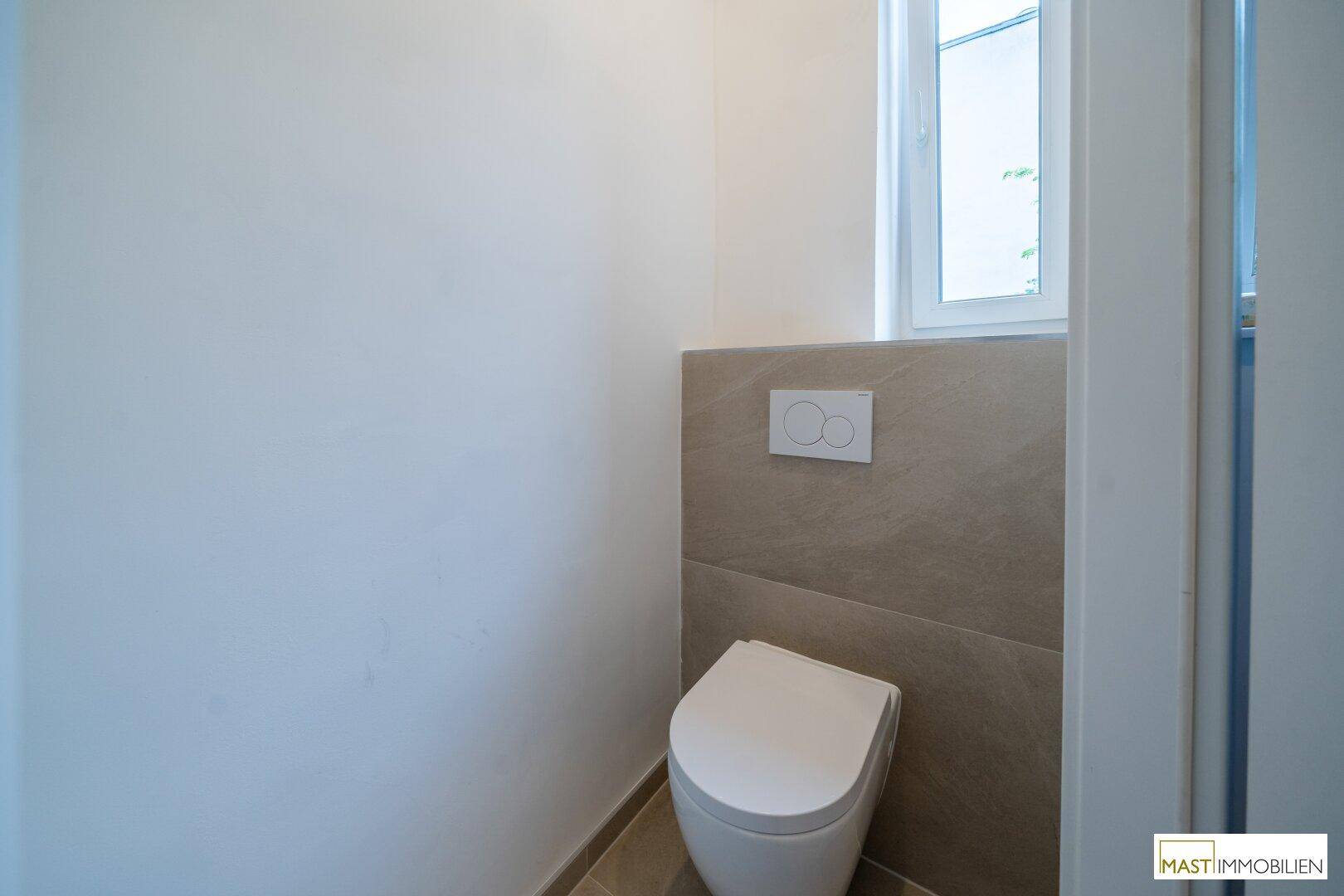 Separate Toilette mit wandhängendem WC, kleinem Fenster und modernen Fliesen.