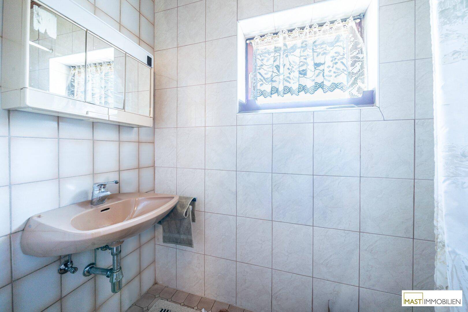 Badezimmer mit hellen Fliesen, einem Waschbecken und einem Spiegelschrank über dem Waschbecken.