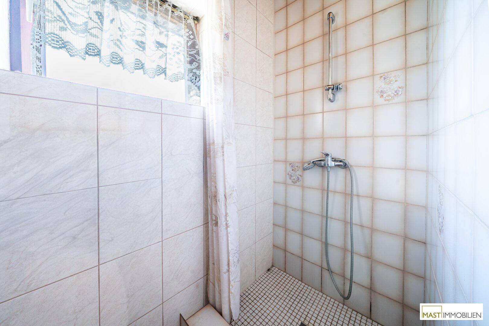 Duschbereich im Badezimmer mit hellen Fliesen und einem Fenster für natürliche Belüftung.
