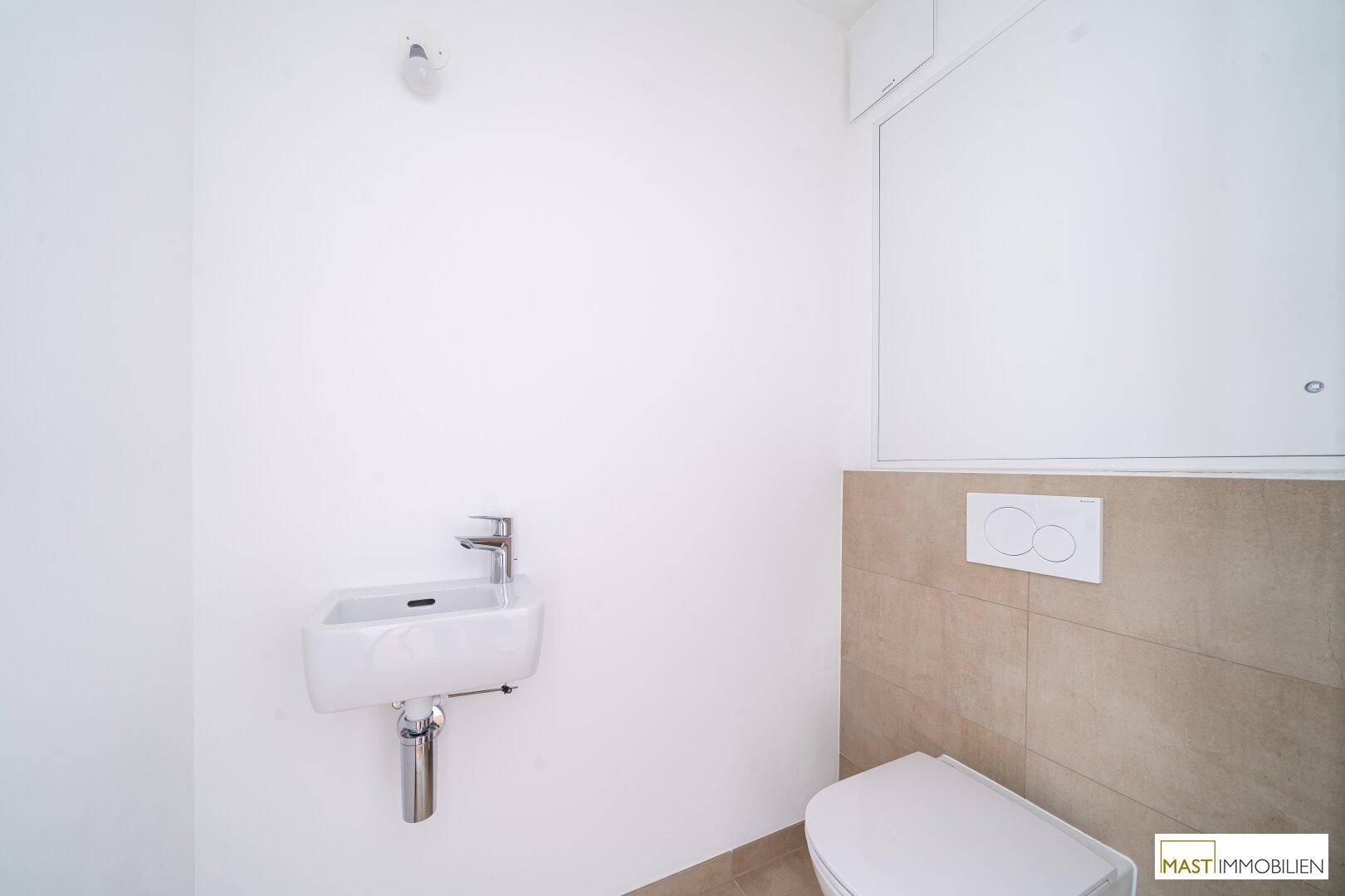 Kleines Gäste-WC mit wandmontiertem WC, kleinem Waschbecken und beigefarbenen Fliesen.