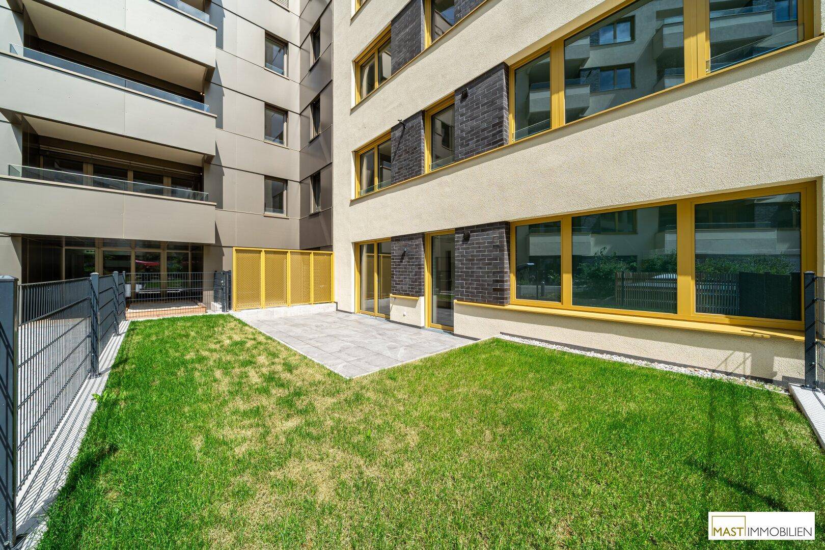 Privater Gartenbereich mit Rasenfläche und kleiner gepflasterter Terrasse vor dem Gebäude.