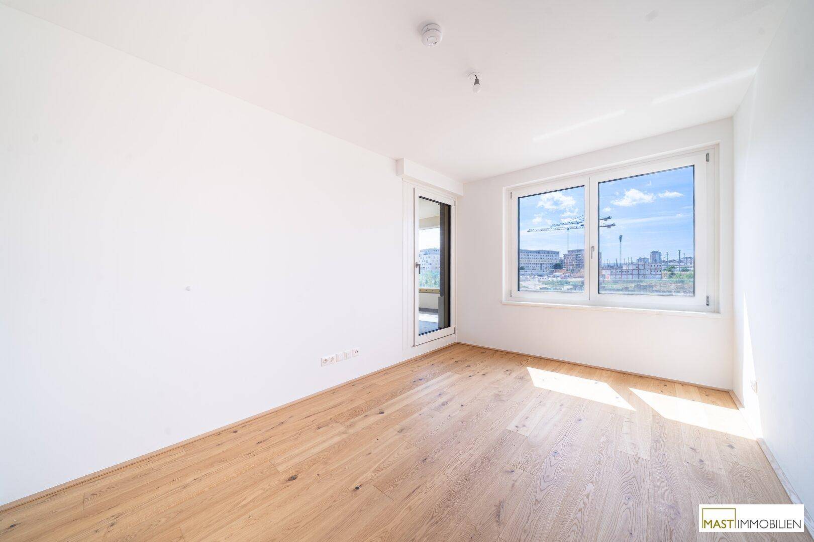 Helles Zimmer mit Holzboden und großem Fenster, bietet Zugang zu einem Außenbereich und viel Tageslicht.