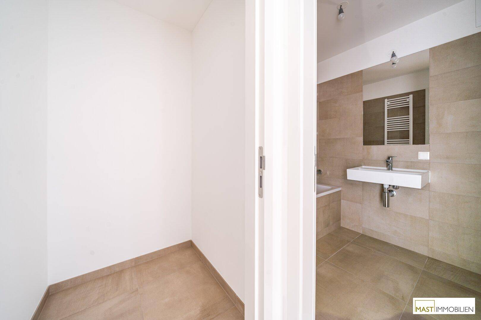 Blick in ein modernes Badezimmer mit beigefarbenen Fliesen und angrenzendem Abstellraum.