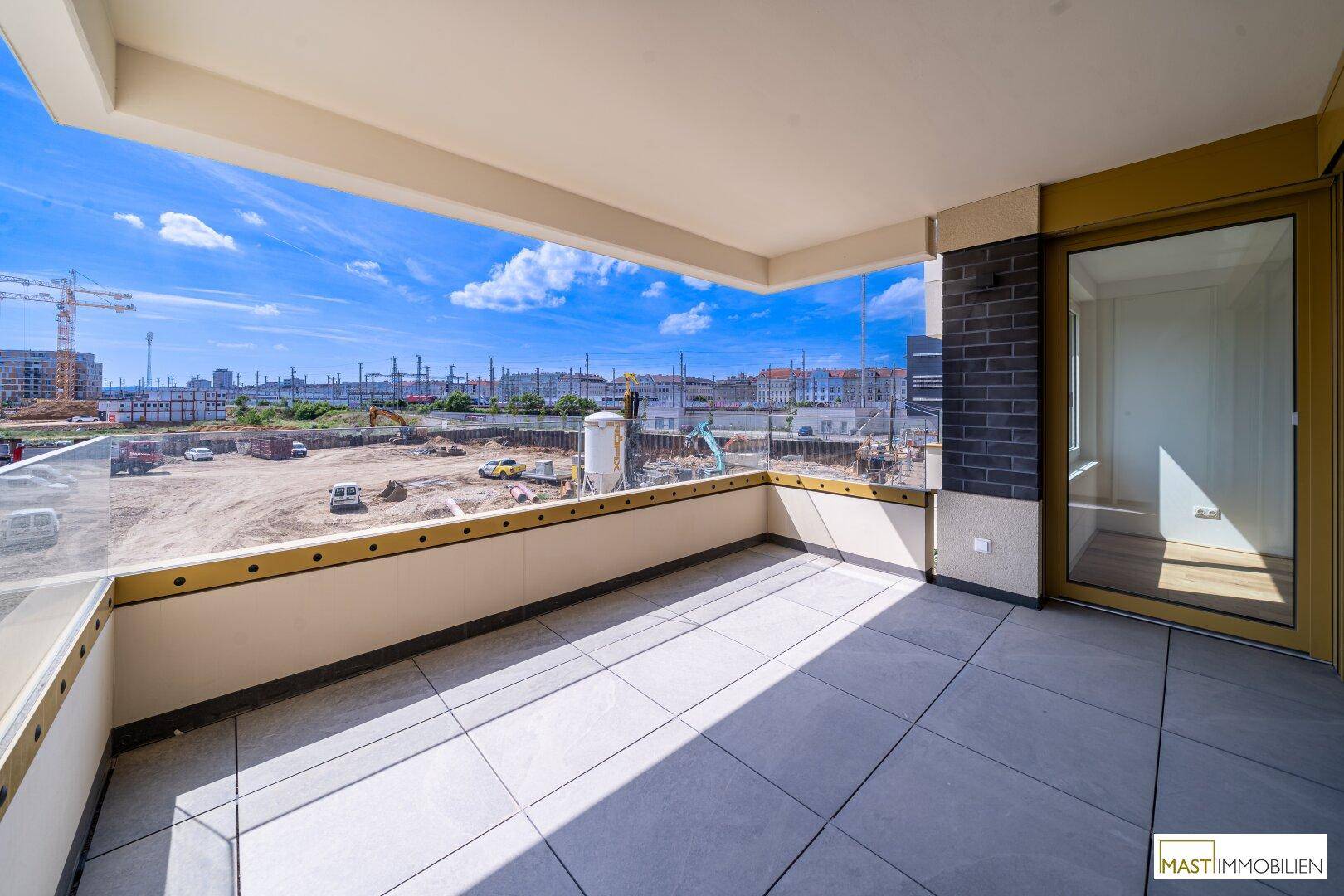 Großer Balkon mit Fliesenboden und Glasbrüstung, bietet weiten Blick auf eine Baustelle und die Stadt.