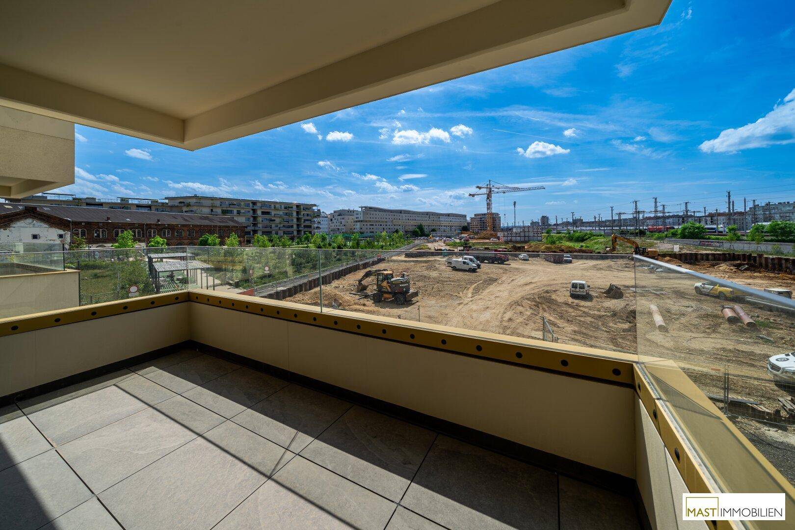 Geräumiger Balkon mit Fliesenboden und Glasgeländer, bietet Blick auf eine Baustelle und die Umgebung.