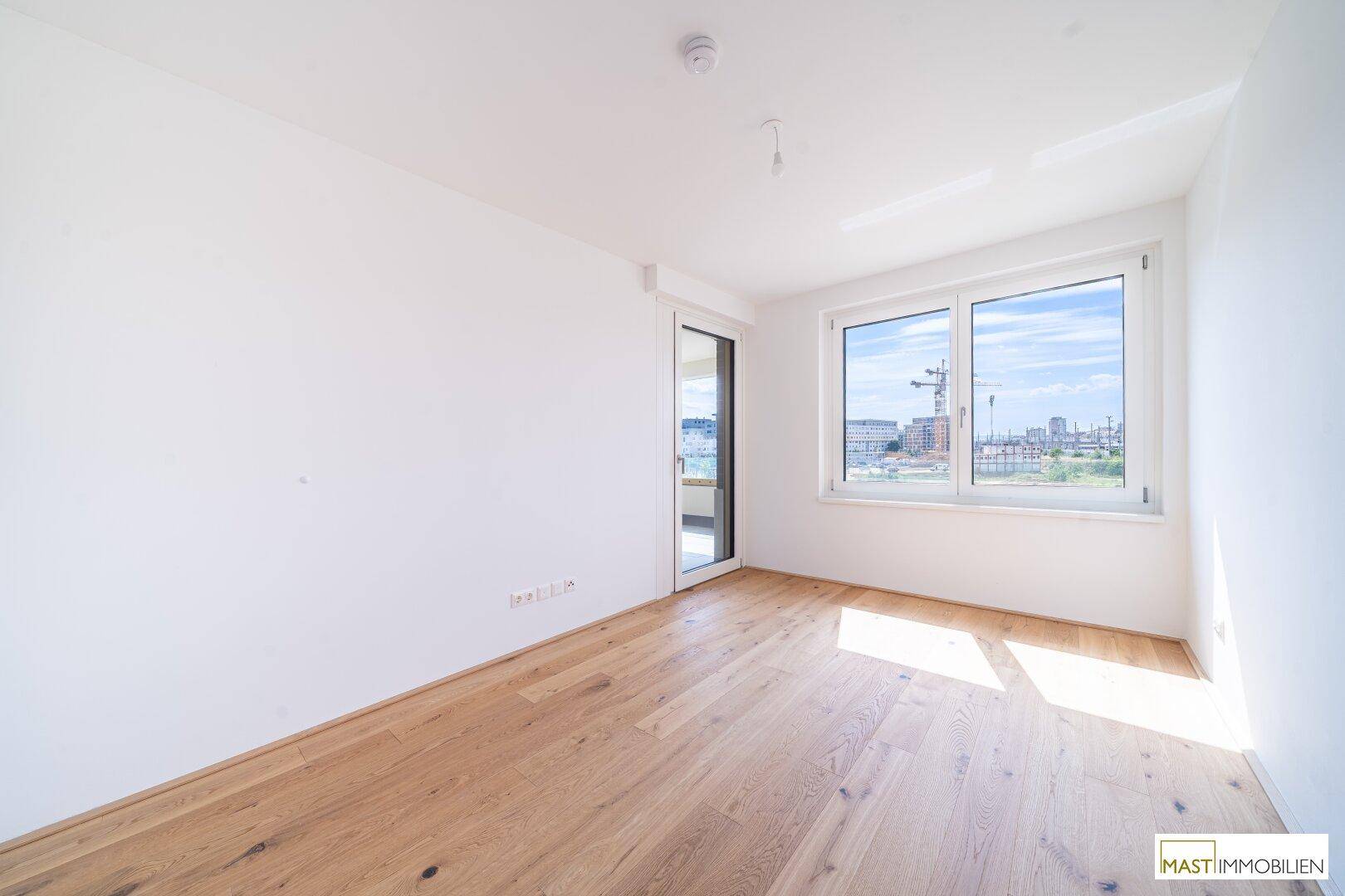 Helles Zimmer mit Holzboden und großen Fenstern, die viel Tageslicht hereinlassen und einen weiten Blick bieten.