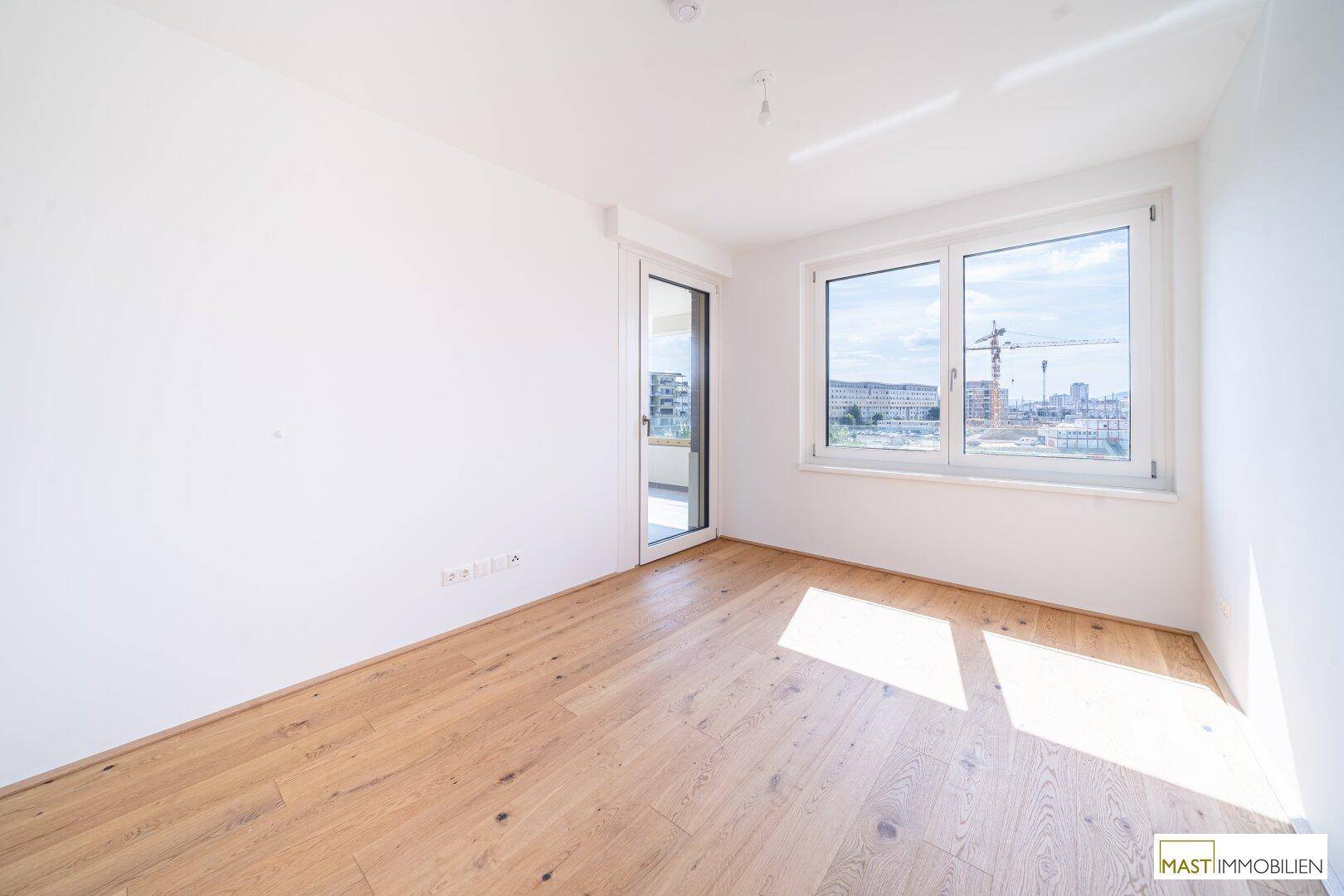 Lichtdurchflutetes, leeres Zimmer mit Holzboden und großem Fenster, ideal für ein Schlafzimmer.