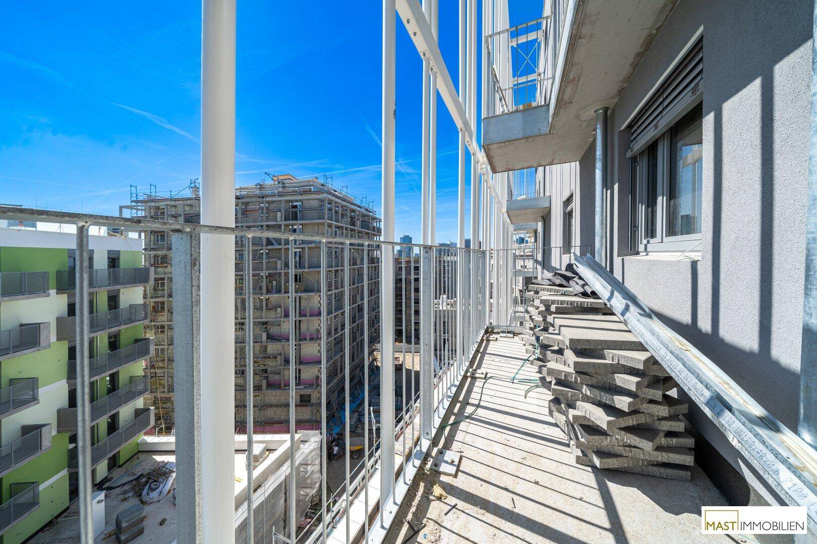 Balkon eines Neubaus mit Blick auf angrenzende Baustellen und moderne Wohngebäude unter blauem Himmel.