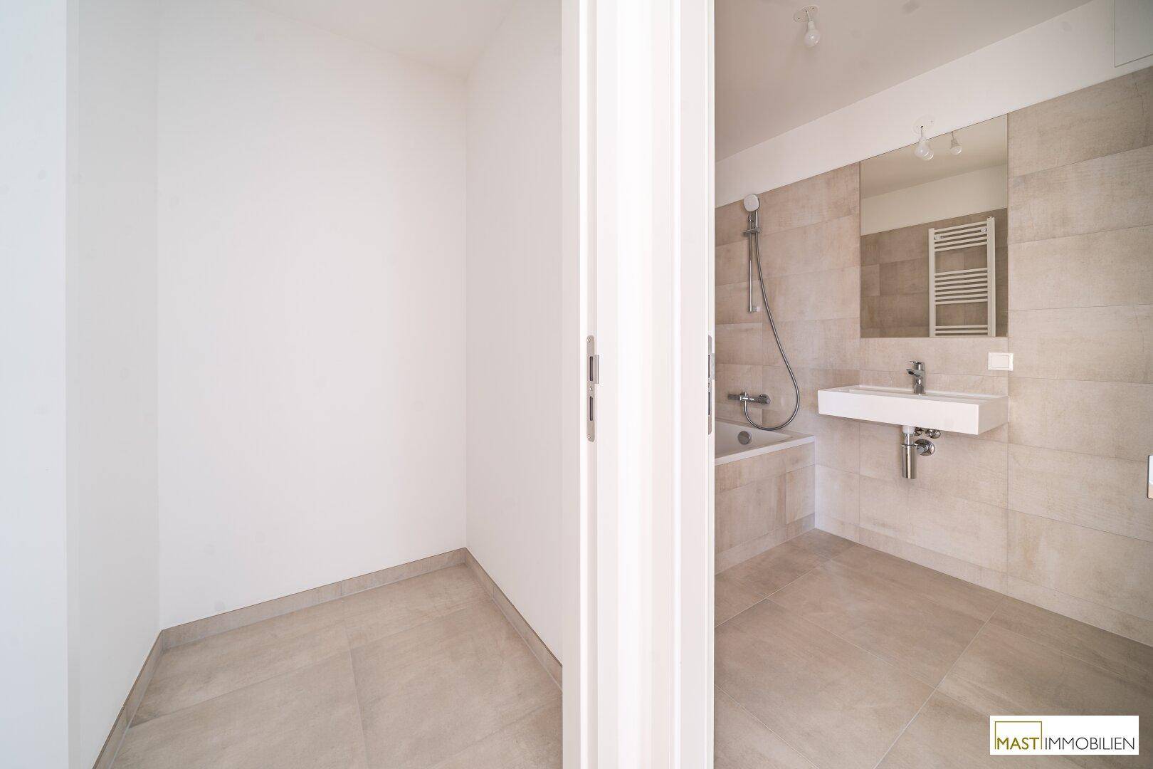 Modernes Badezimmer mit hellen Fliesen, Badewanne und Waschbecken, daneben ein praktischer Abstellbereich.